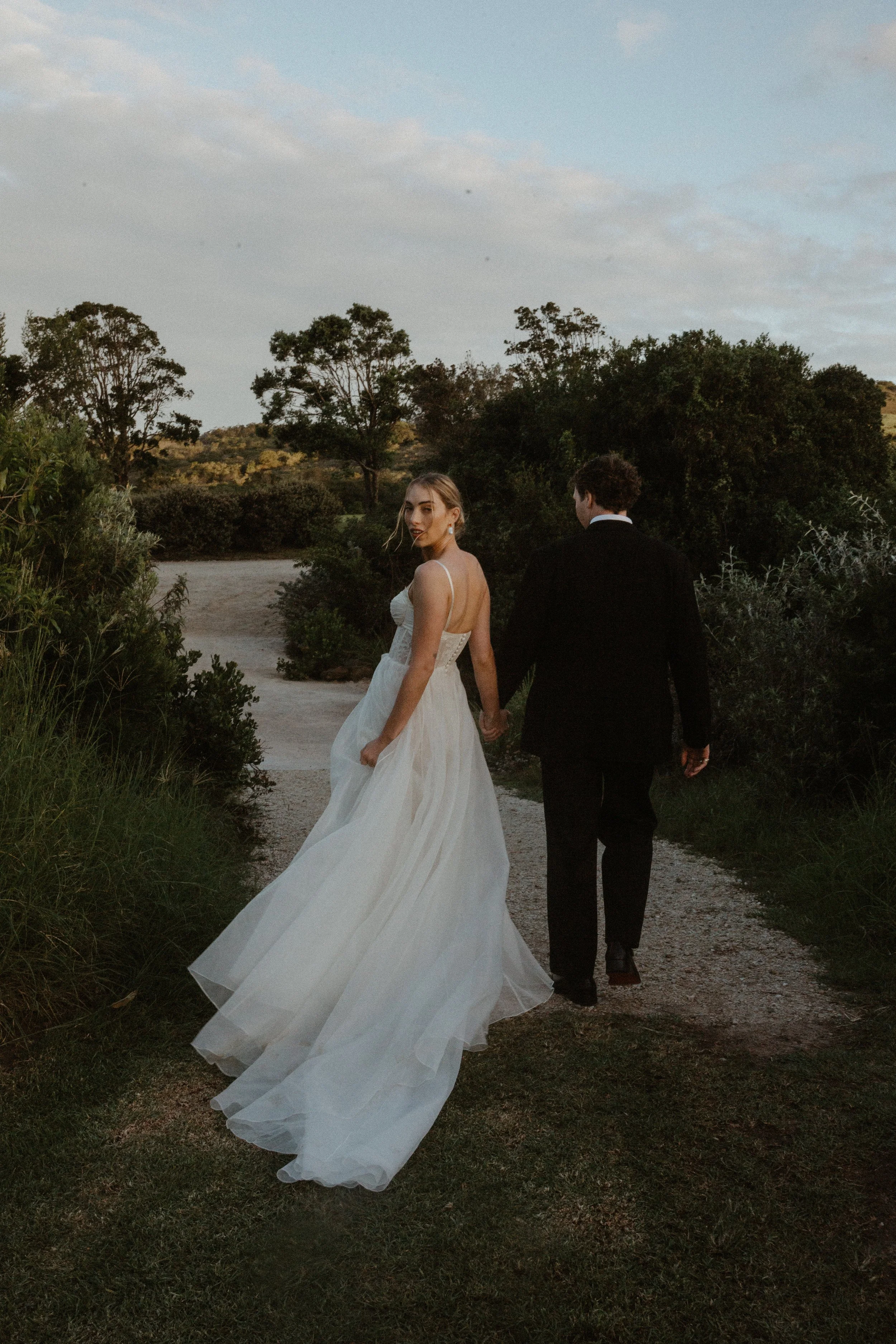 A bride and groom walking outdoors on a dirt path in a rural setting, holding hands, with trees and hills in the background, during the early evening.