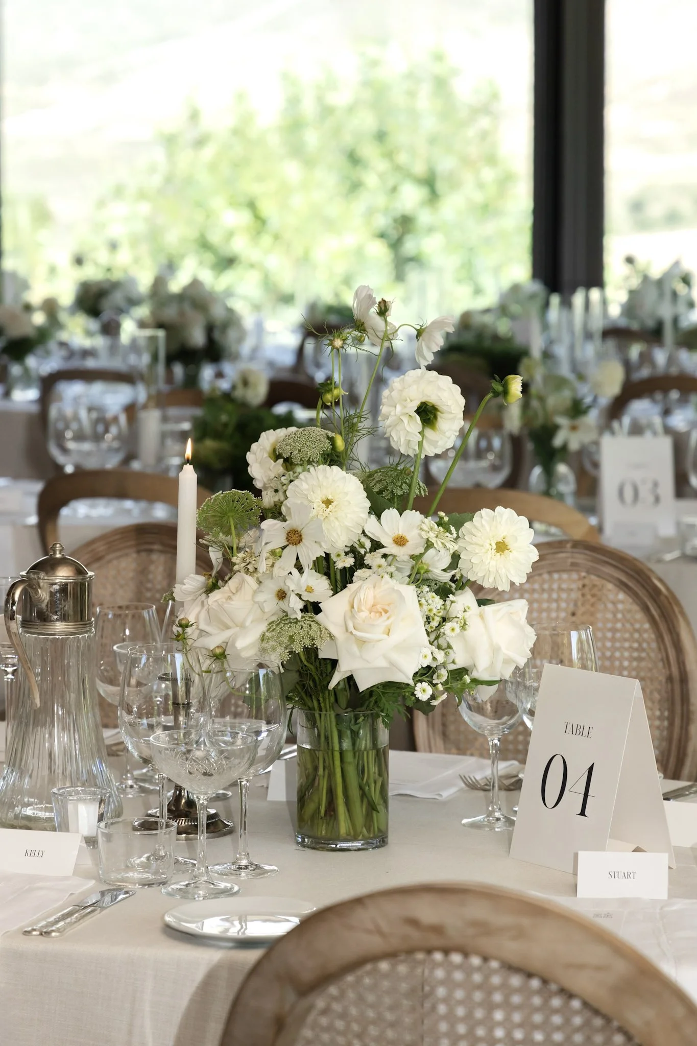 A floral centerpiece of white flowers and greenery on a dining table at an elegant event with table numbers and glassware.