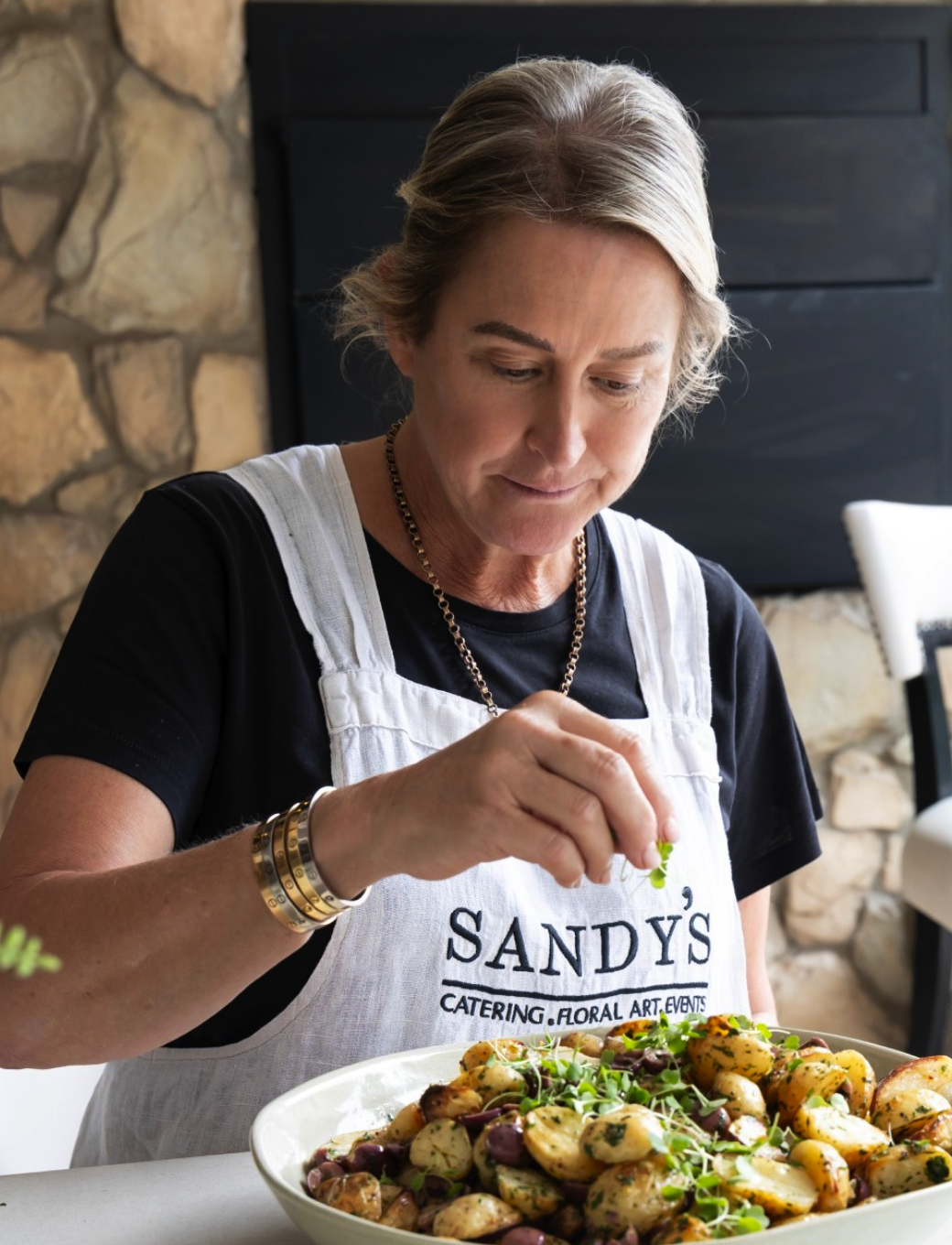 A woman wearing a white apron with 'SANDY'S' embroidered on it is garnishing a large bowl of roasted potatoes with greens in a kitchen setting.