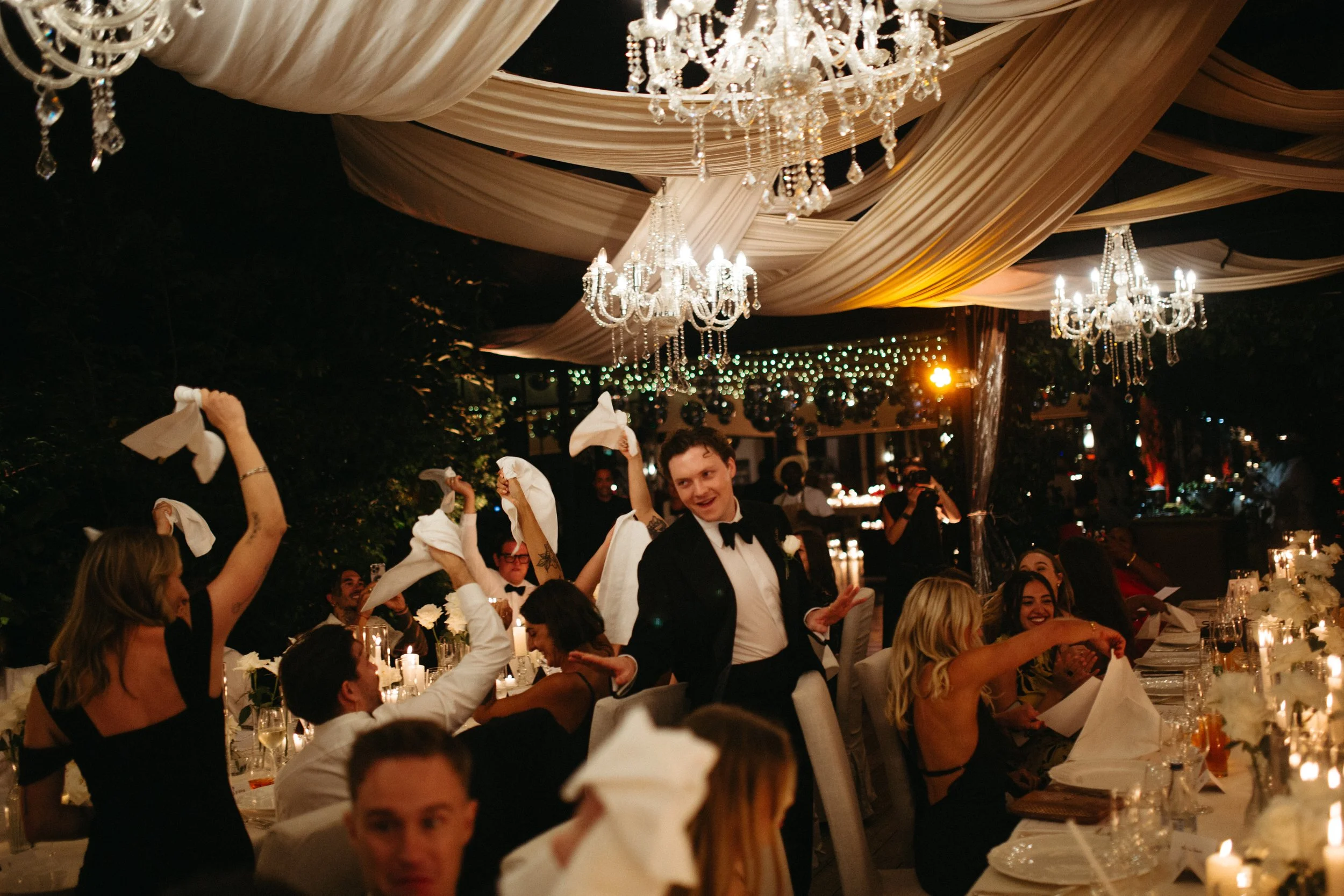 A festive celebration at night with chandeliers and draped fabric decorations. Guests are seated at elegant tables with candles, and some guests are waving napkins in the air. A waiter in a tuxedo is serving or dancing among the guests, smiling and e