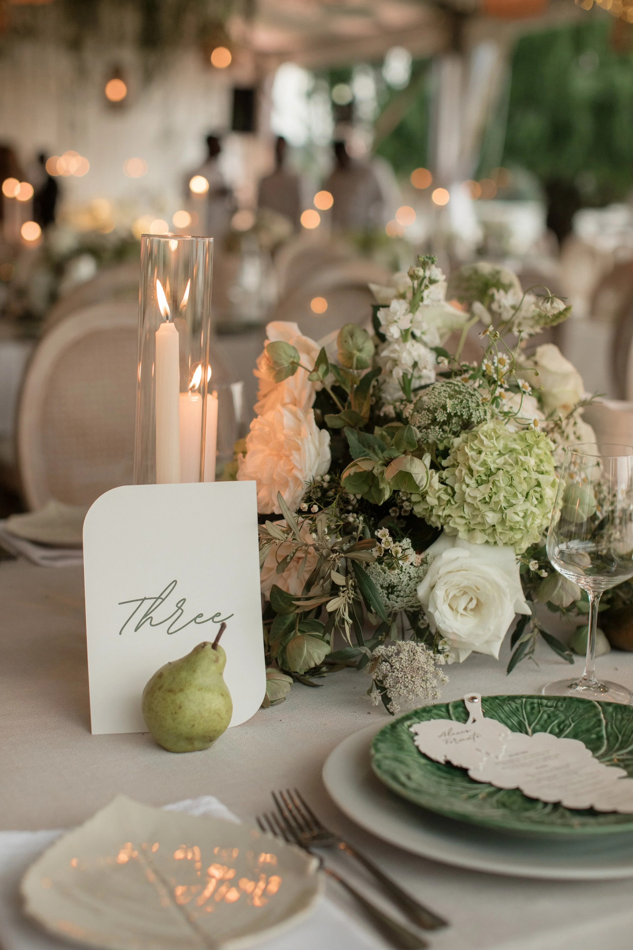 A table setting at a wedding reception featuring a floral centerpiece with white and green flowers, a tall glass candle holder with lit candles, a pear, a place card labeled 'Three', and elegant dinnerware and glassware.