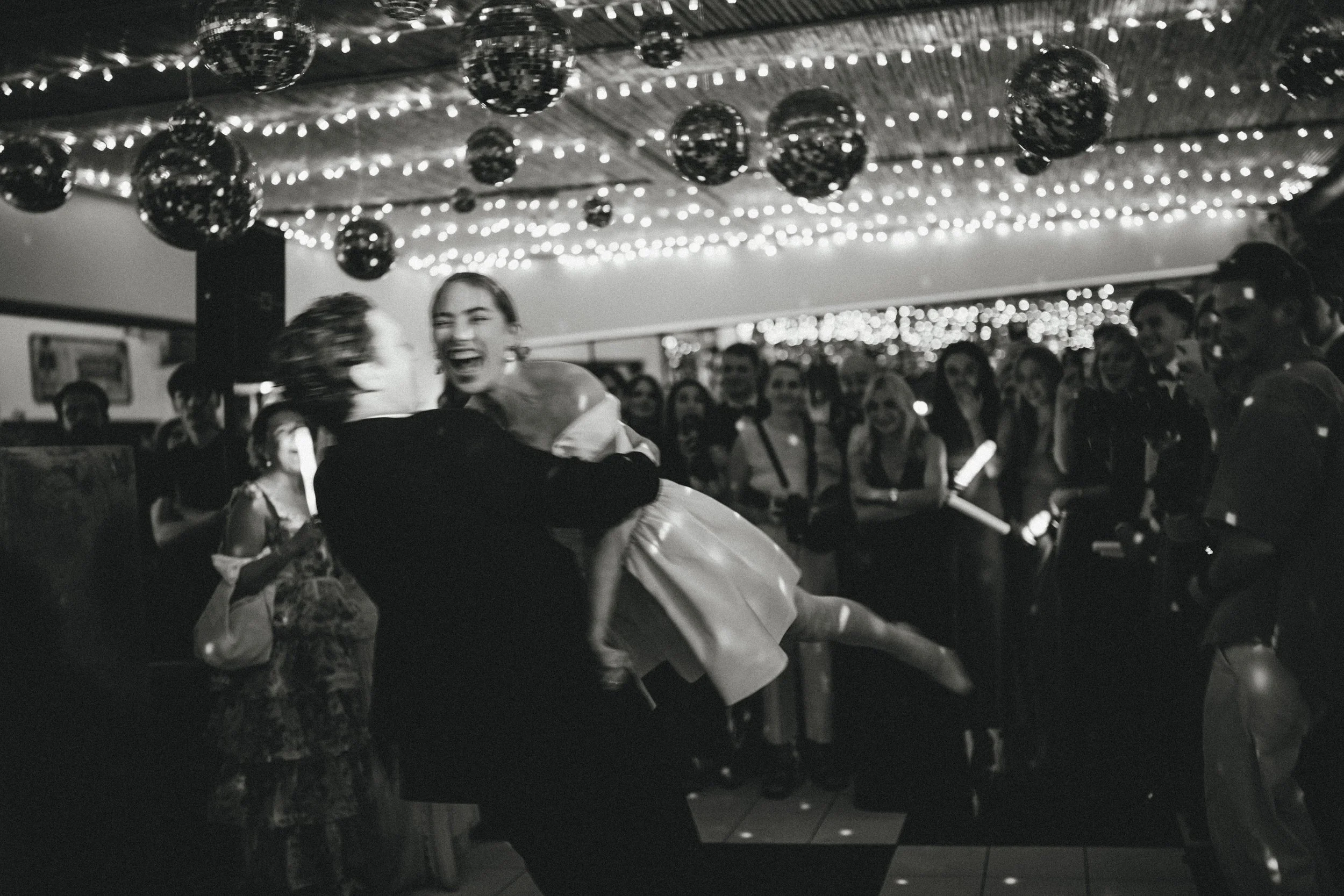 A black and white photo of a celebration or dance event. Two women are dancing closely in the foreground, laughing. One wears a floral dress, the other a white dress. In the background, there are several people watching, and hanging string lights and