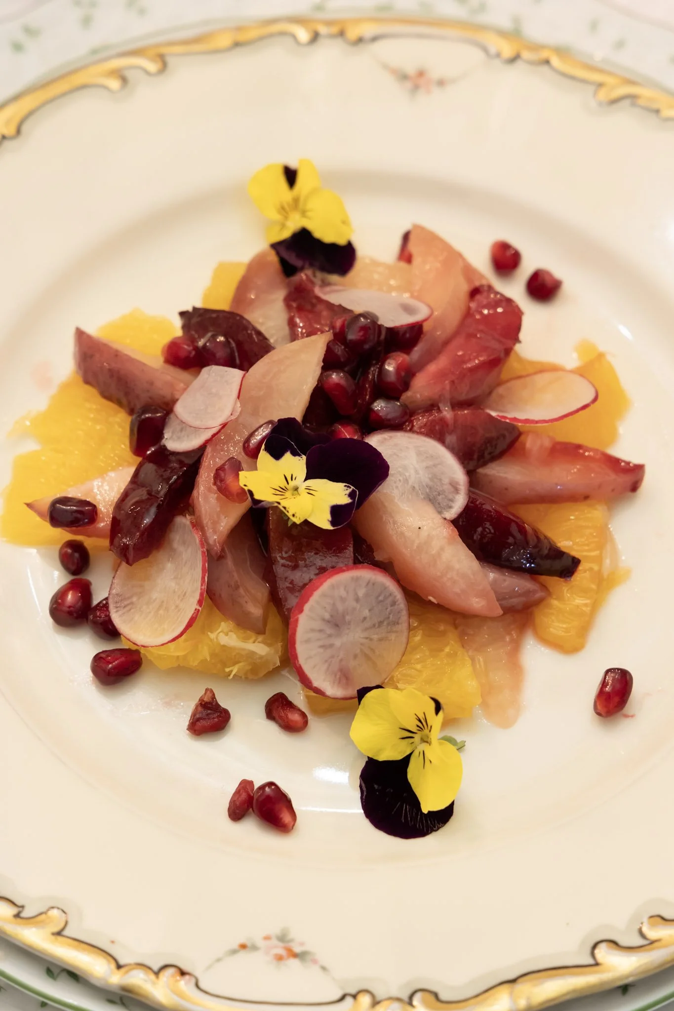 A plated dish featuring sliced bird meat garnished with pomegranate seeds, radish slices, orange segments, and edible flowers on a porcelain dish.