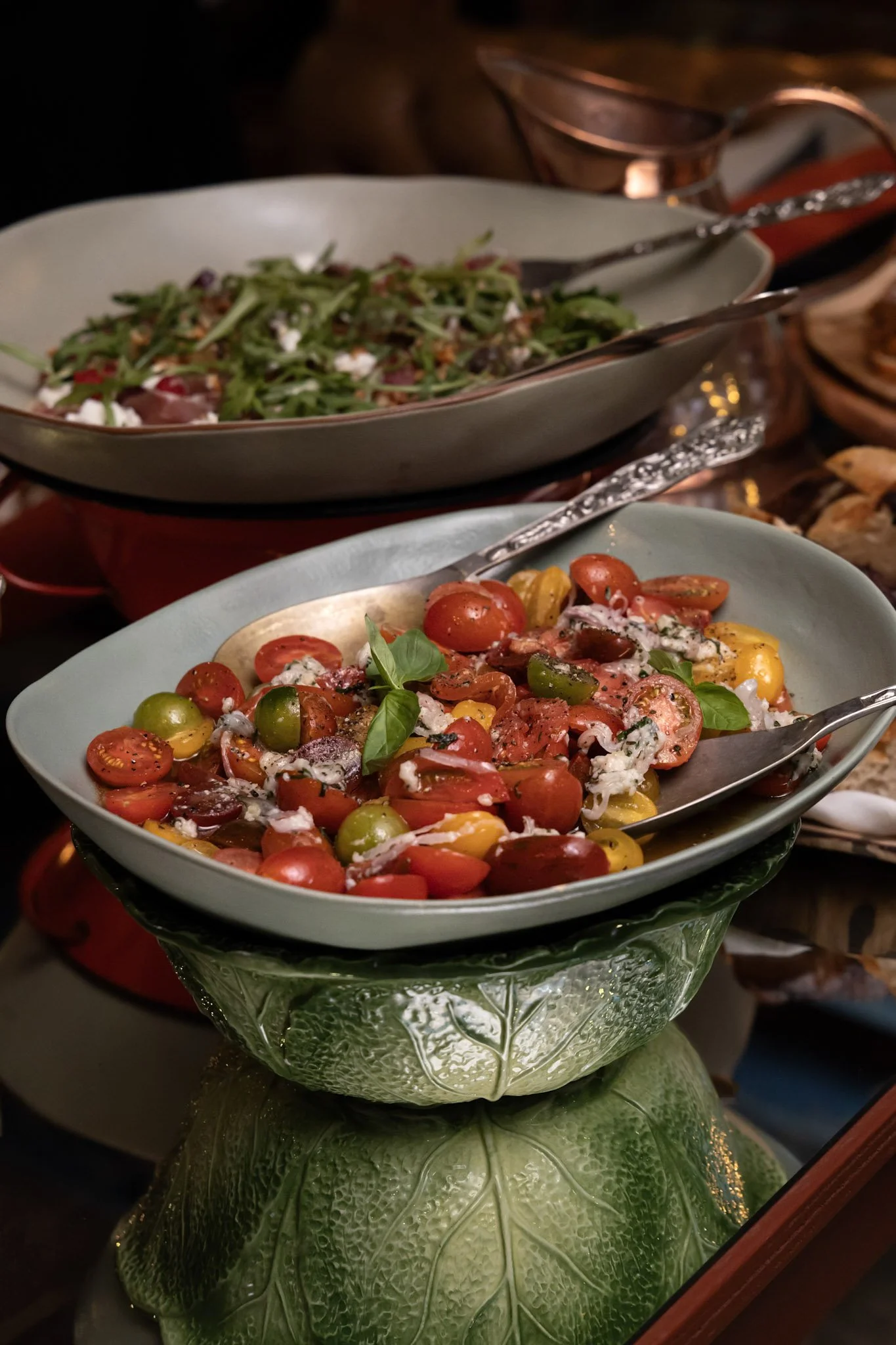 A bowl of colorful cherry tomato salad with basil, served in a decorative glass bowl, on a table with other dishes and serving utensils in the background.