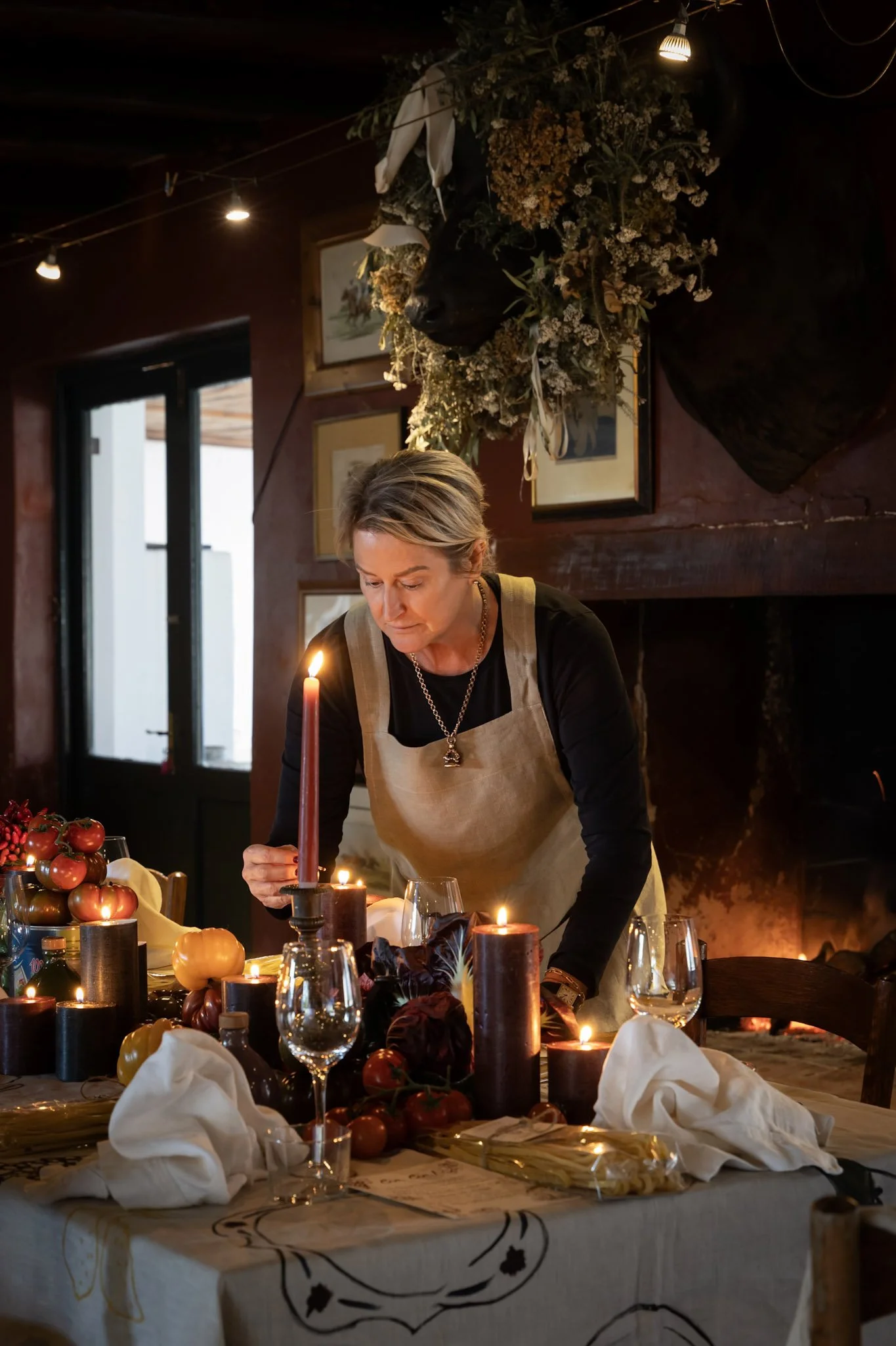 A woman in a beige apron decorates a table with candles, pumpkins, and fall-themed decorations in a cozy indoor setting, with a large floral and animal head wall hanging in the background.