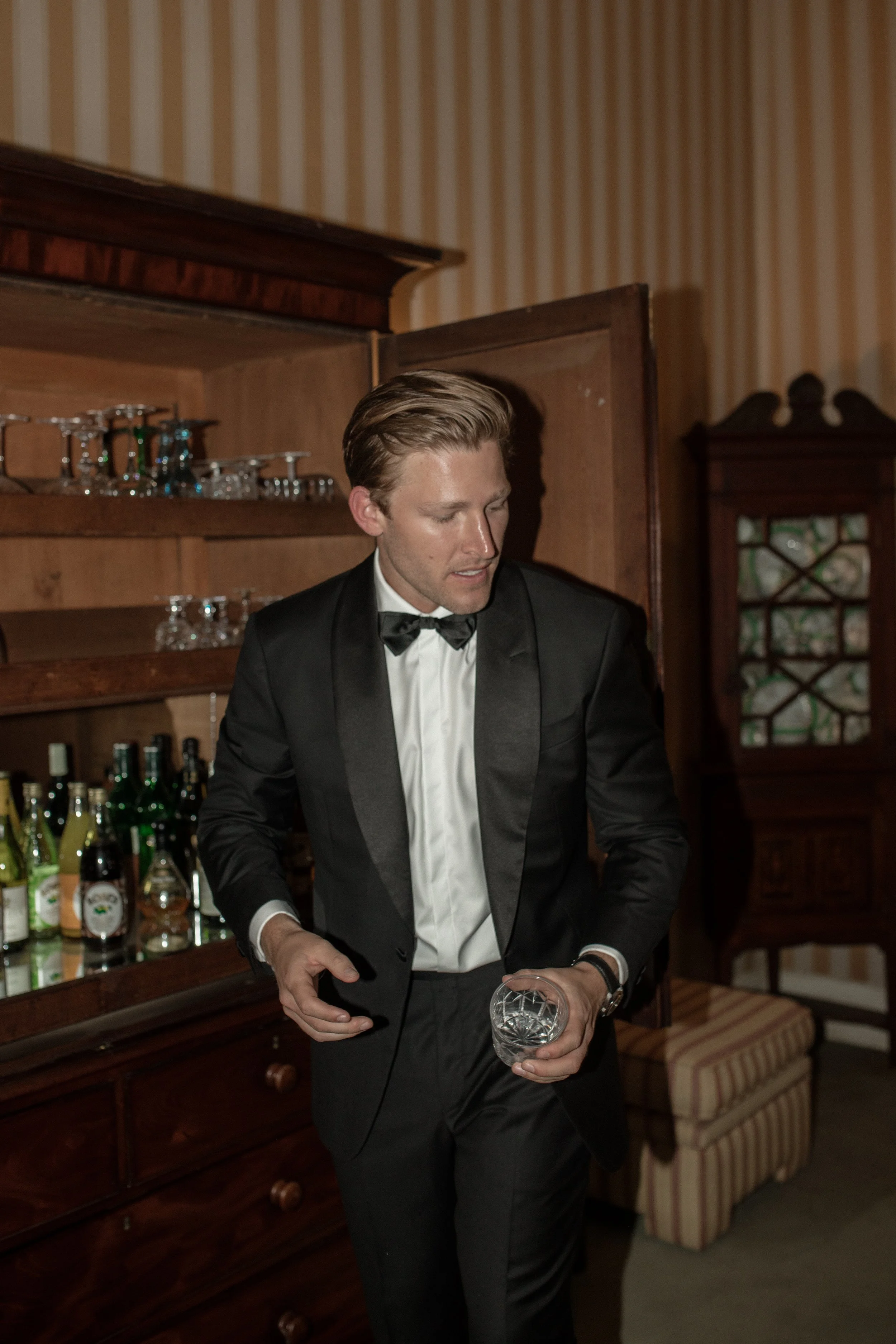 A man in a tuxedo pouring a drink in a room with wooden shelves and barware.