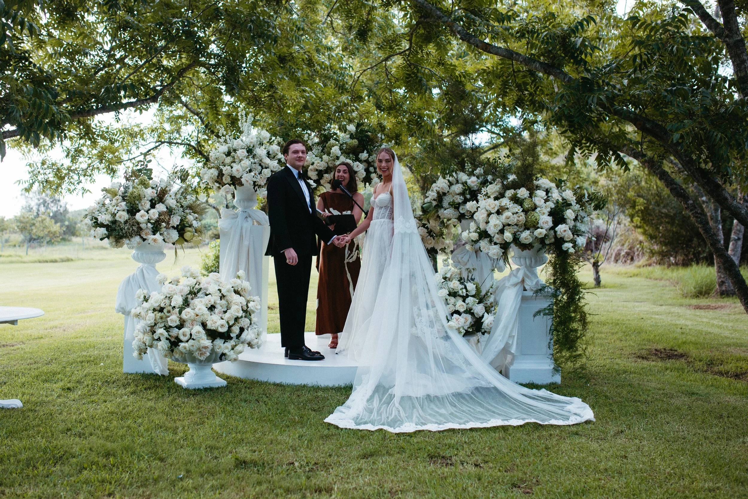 A bride and groom exchanging vows during an outdoor wedding ceremony under a large tree, surrounded by floral arrangements.