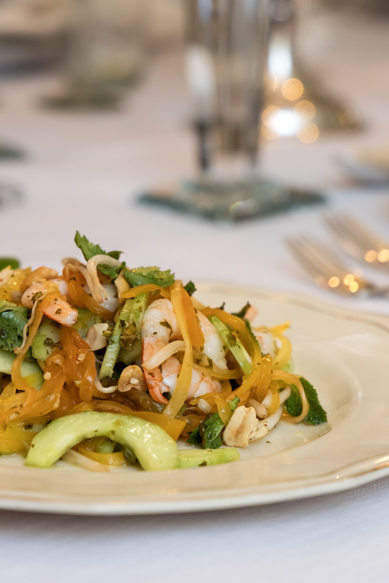 Close-up of a colorful shrimp and vegetable noodle salad on a white plate with a blurred background of a dining table and silverware.