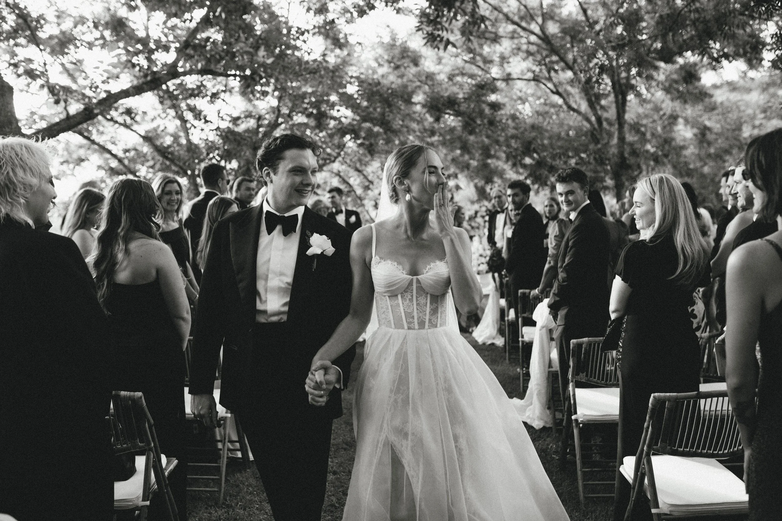Bride and groom holding hands at an outdoor wedding ceremony, surrounded by seated guests in formal attire, under trees with sunlight filtering through the leaves.