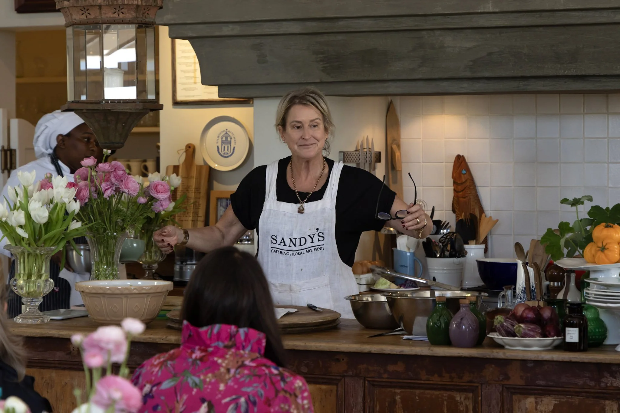 A woman wearing a white apron with the words "SANDY'S CATERING, FLORAL, ART, EVENTS" offers a demonstration in a kitchen with children watching. The kitchen counter is filled with flowers, vegetables, and kitchen utensils.