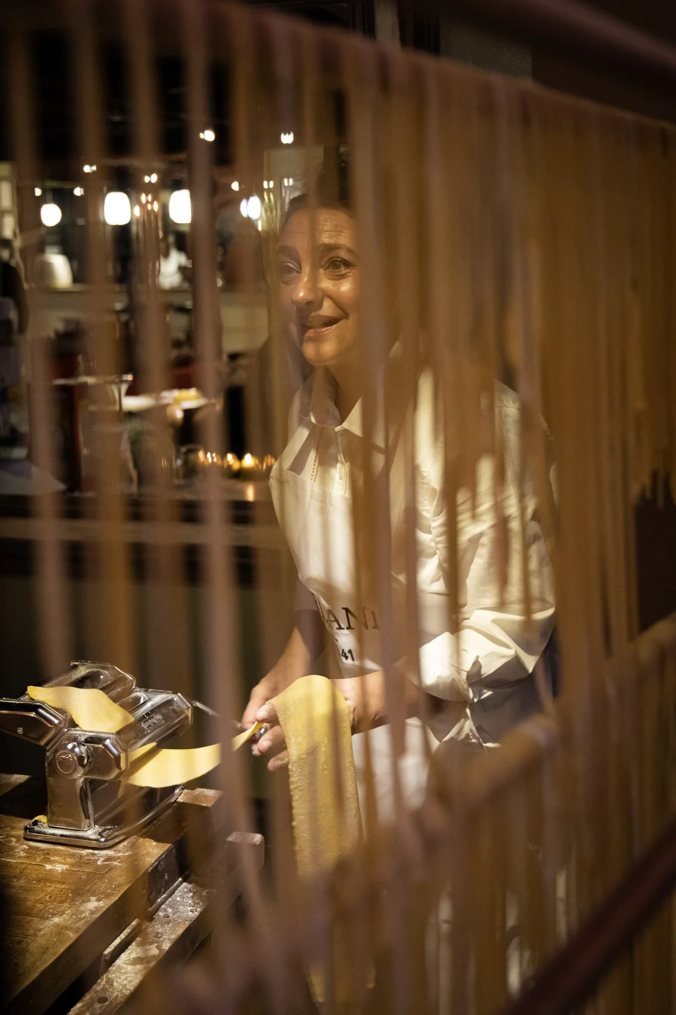 A woman in a white apron is slicing pasta at a pasta machine in a restaurant kitchen, seen through a wooden lattice partition.