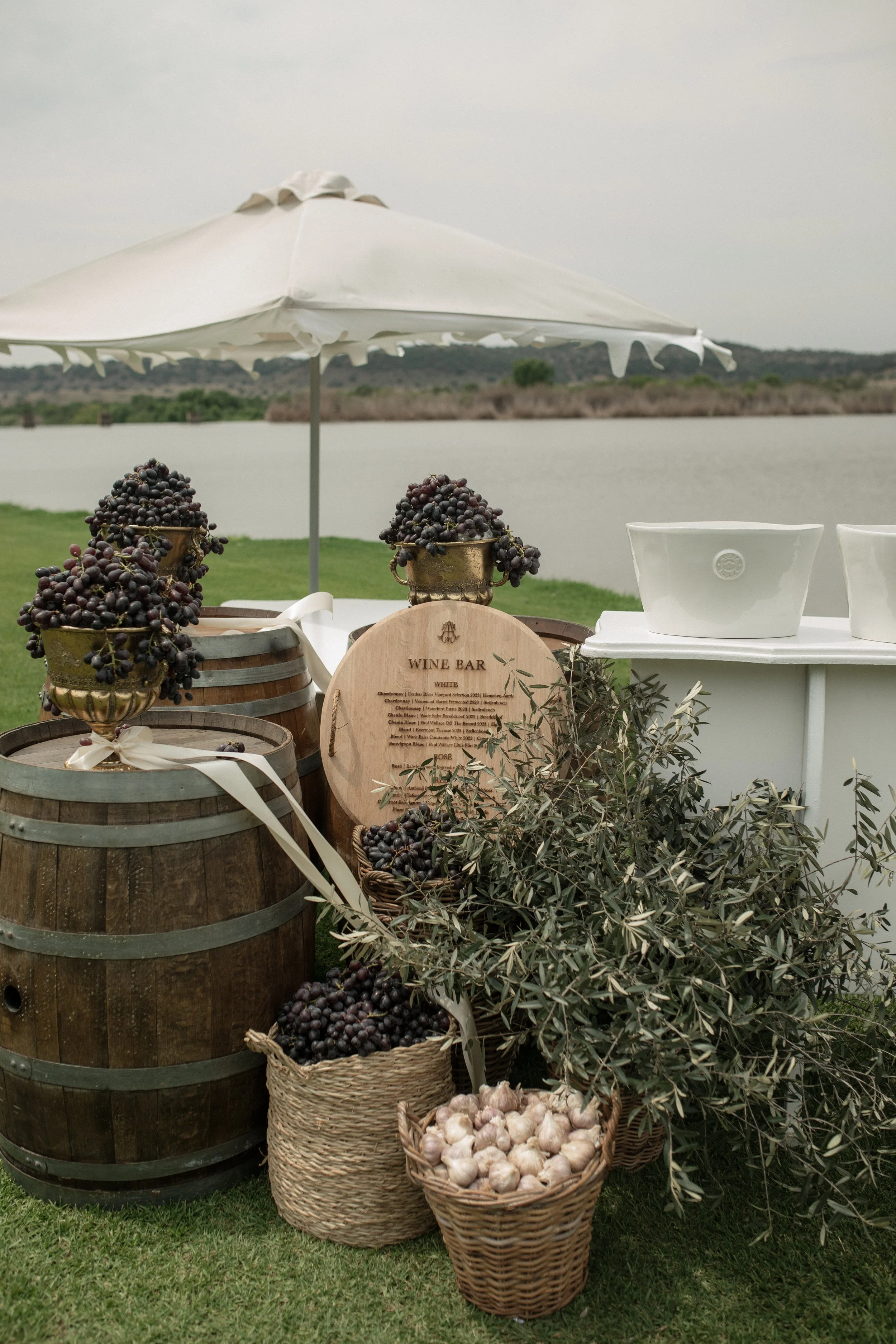 A wine bar setup near a body of water with barrels, baskets of garlic, olive branches, and clusters of grapes, under a white patio umbrella.