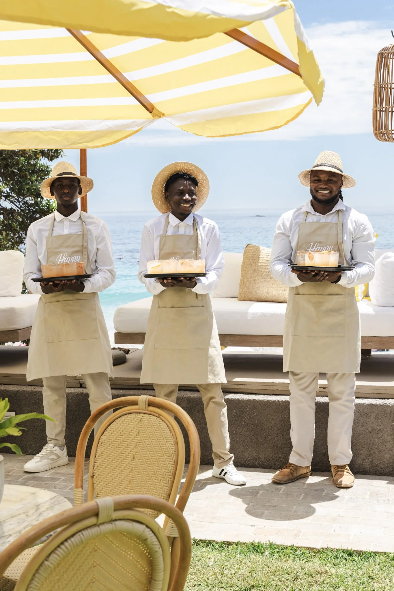 Three male waiters dressed in white shirts, beige aprons, and straw hats are standing outdoors under a yellow and white striped umbrella at a beachside restaurant. They are each holding trays with a dessert that has the word 'Happy' on it, and they a