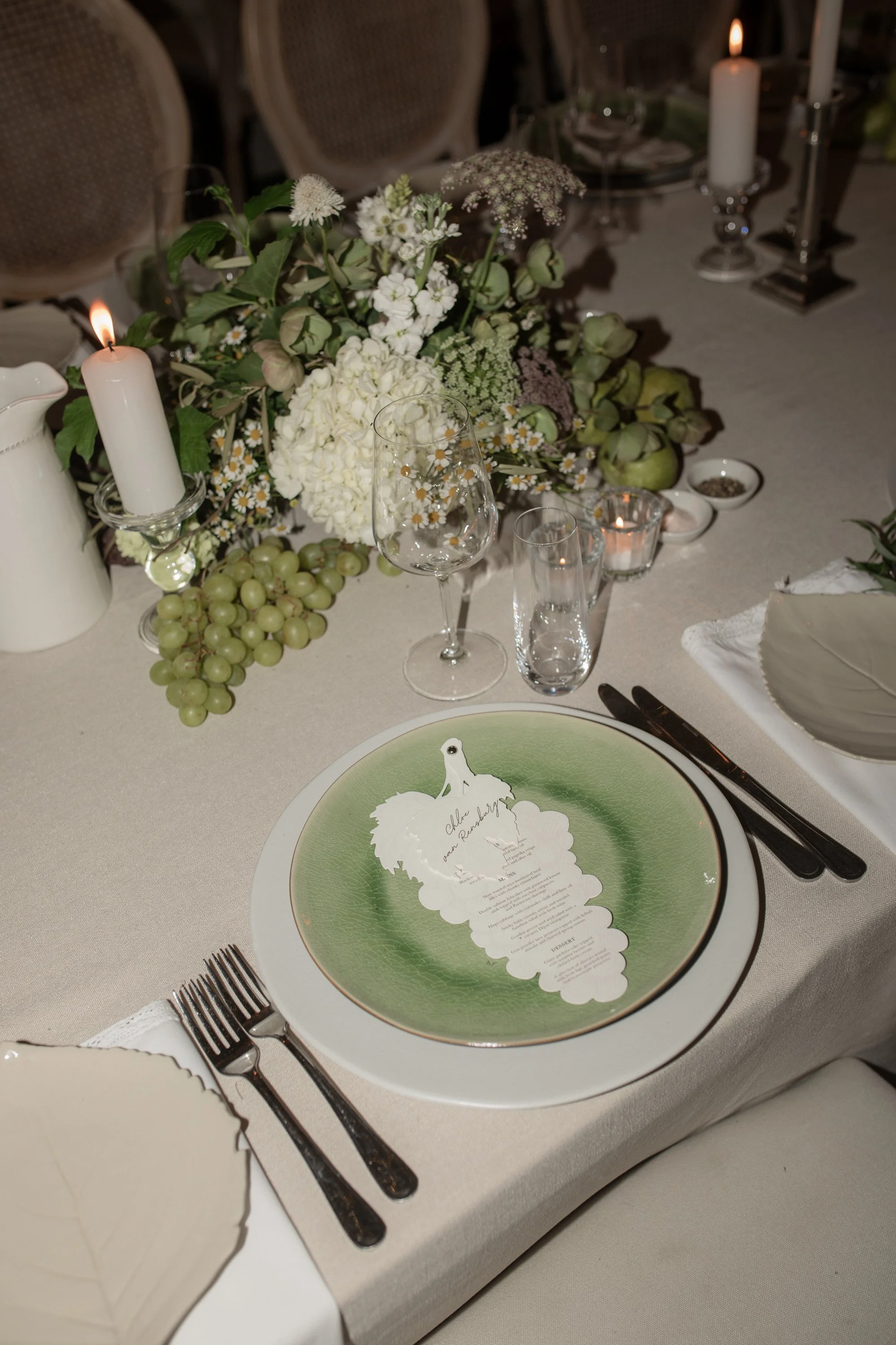 A beautifully decorated table with a floral centerpiece, white candles, glassware, and a plate with a menu and a cut-out of a bride and groom, likely at a wedding reception.
