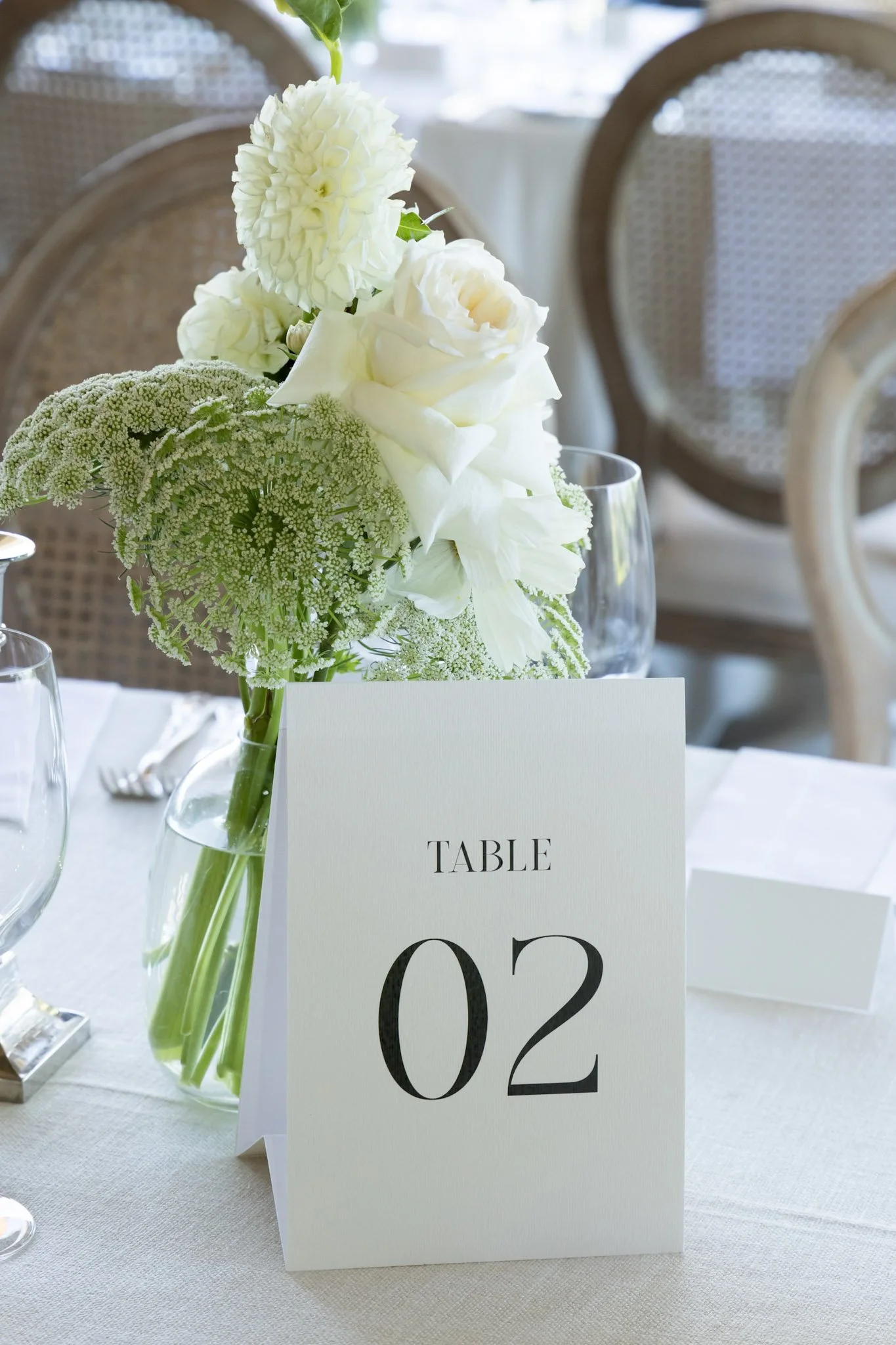 A floral centerpiece with white flowers in a glass vase on a table, with a table number card reading '02' and a sign with the word 'TABLE' in a formal setting.