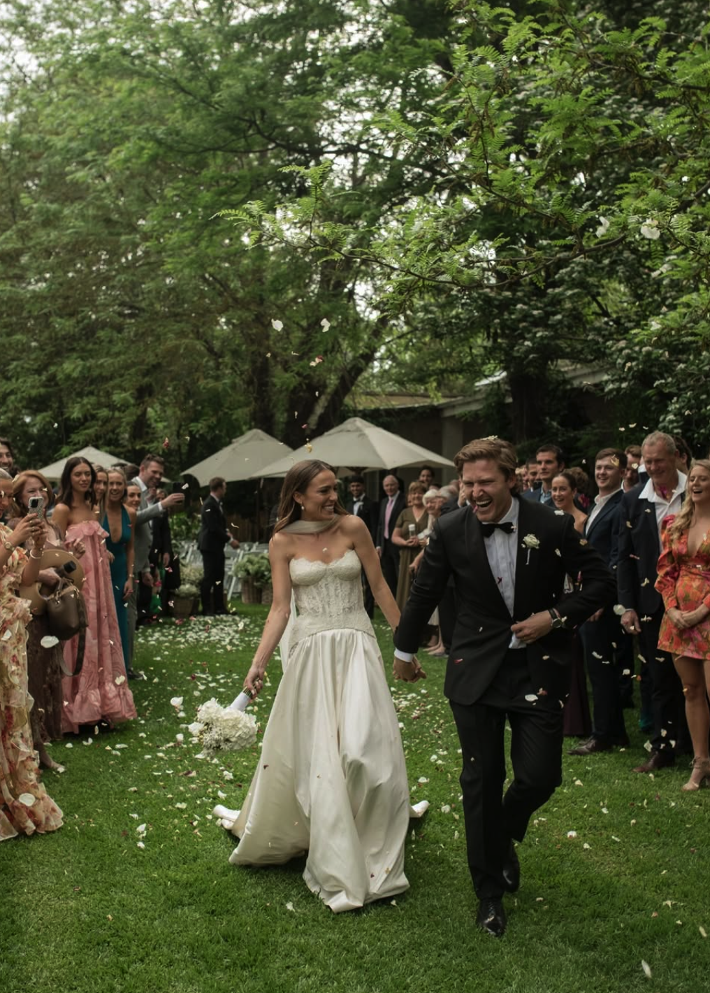 A newlywed couple walking hand-in-hand through a garden with family and friends, celebrating their wedding with flower petal throw. The bride is in a white wedding dress, holding a bouquet, and the groom is in a black tuxedo. Guests are smiling and taking photos.