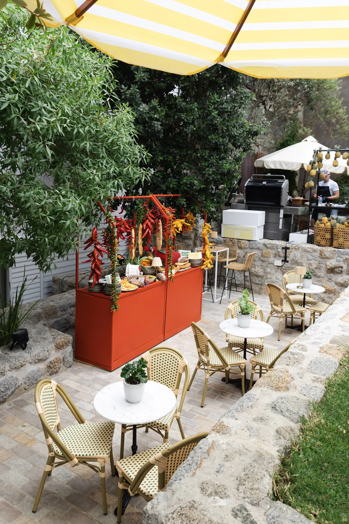 An outdoor patio café setting with white and green patterned chairs and round white tables, potted plants, a red food stand with hanging chili peppers and bread, a grill, and umbrellas for shade. Lush green bushes and trees surround the area.