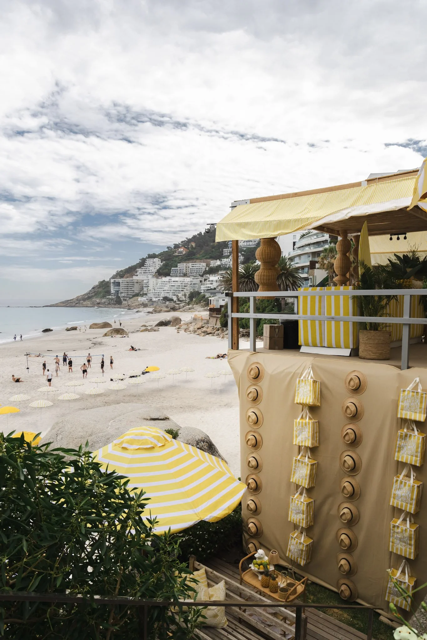 Beach scene with yellow and white striped umbrellas, a beach bar with yellow and white striped decor, and people playing volleyball with white umbrellas on the sandy shore. Buildings and a hillside with houses are in the background under a partly clo