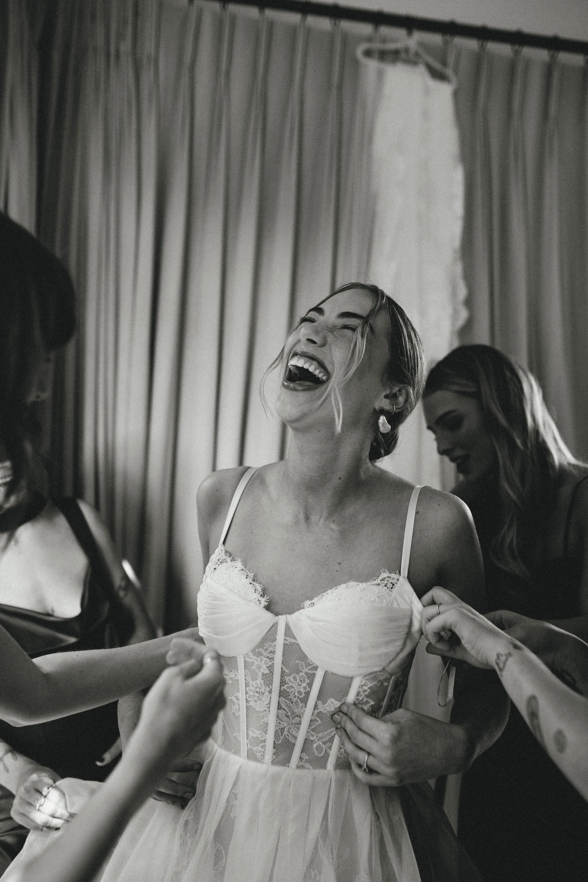 A woman in a wedding dress laughing happily as she gets ready with friends.