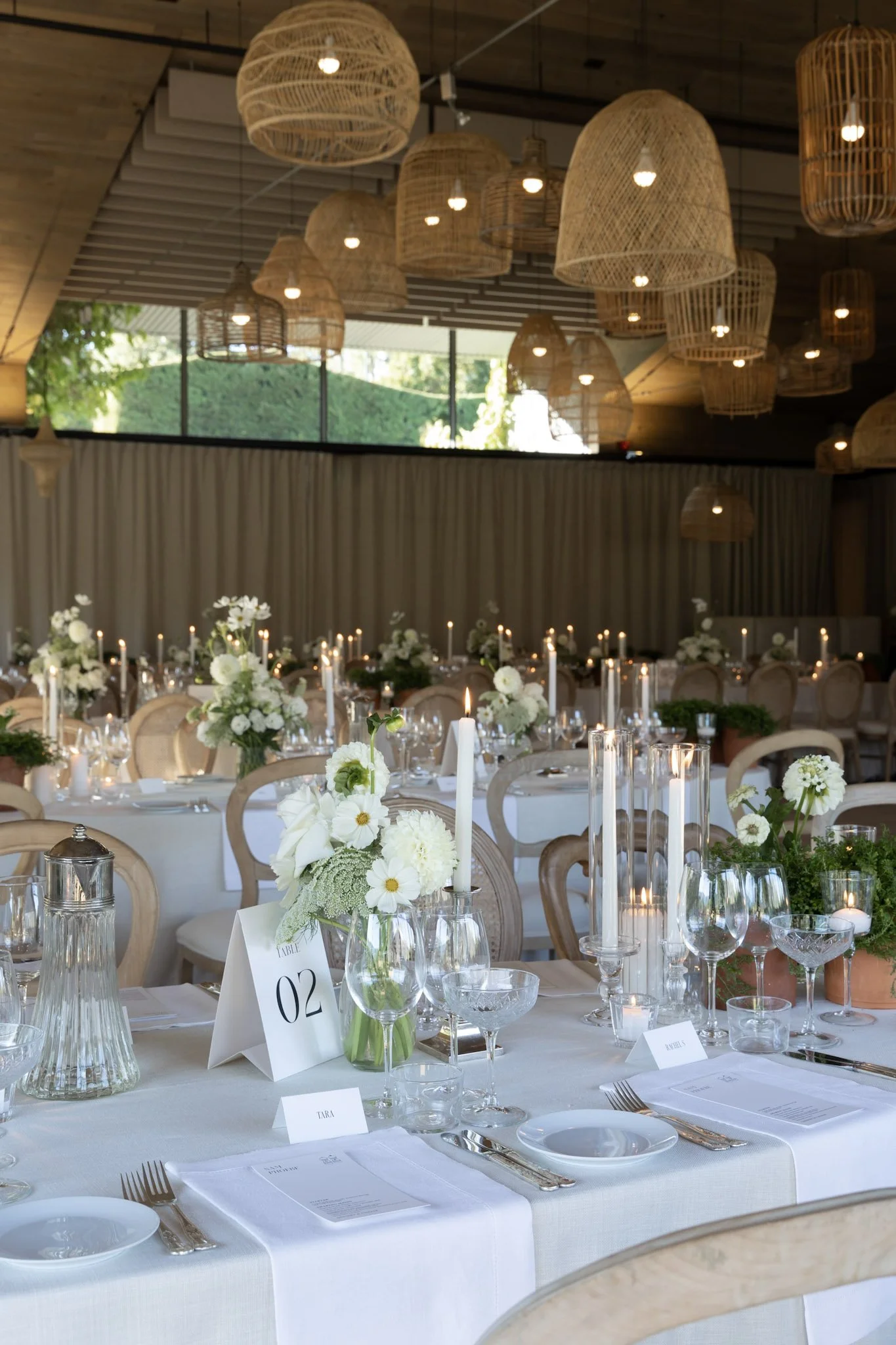 Elegant banquet hall decorated for a wedding reception with white tablecloths, floral centerpieces, and candles, with wicker pendant lights hanging from the ceiling and large windows showing greenery outside.