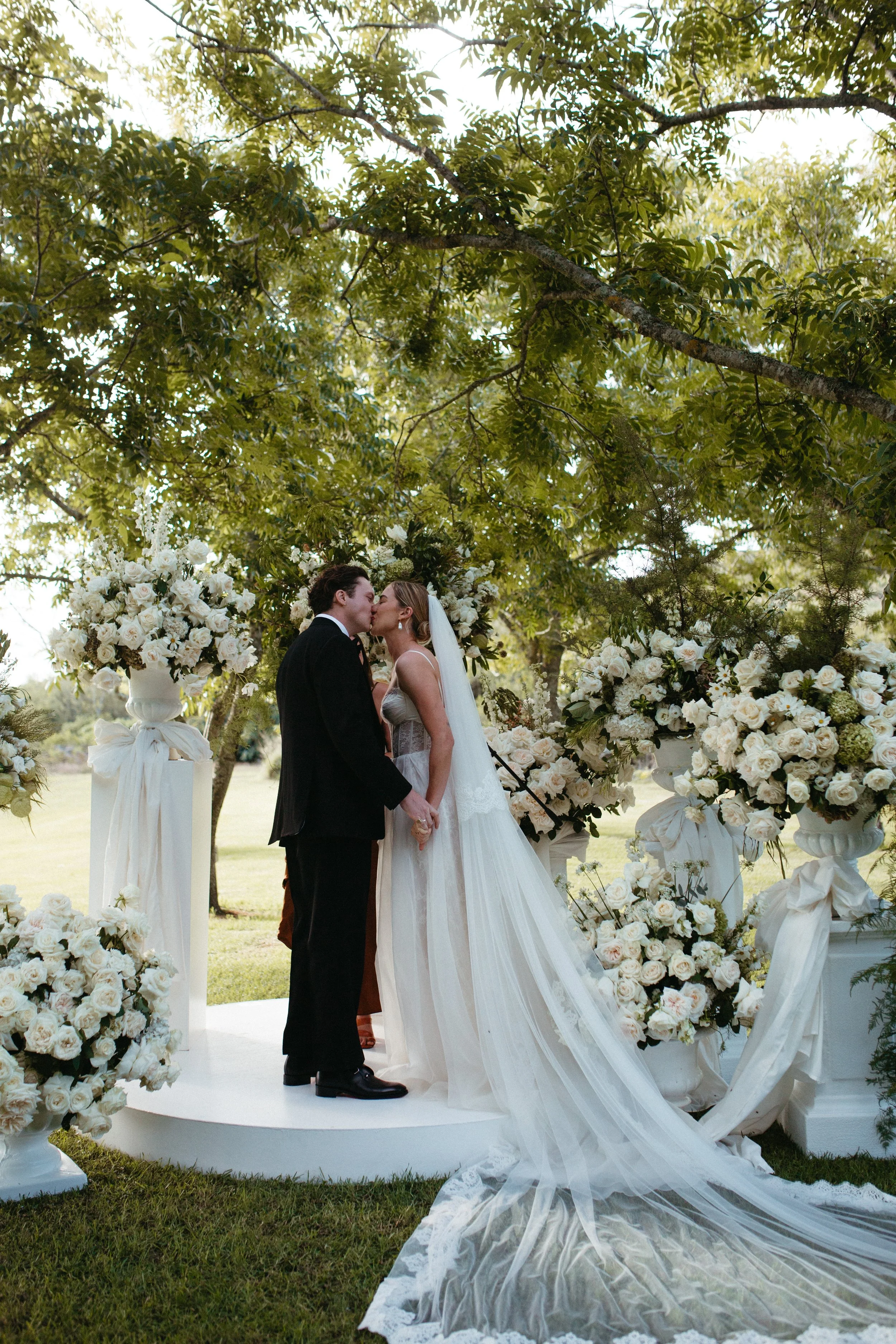 A bride and groom kissing during their outdoor wedding ceremony under a green leafy tree, with floral arrangements in vases around them.