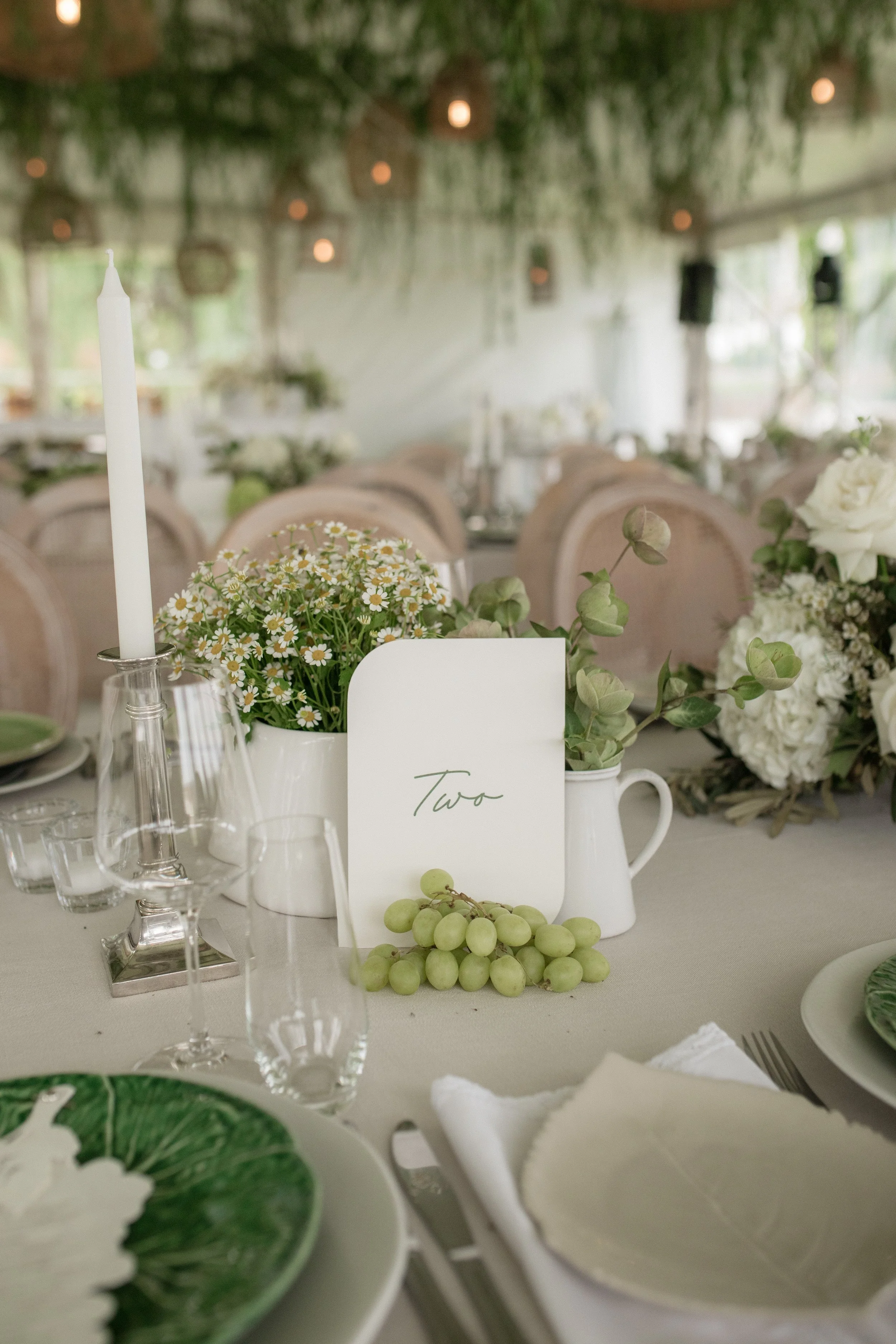 Table decorated with green grapes, a white pitcher with flowers, a white card with cursive writing, surrounded by glasses, plates with green and white food, and a candle. Background shows blurred chairs and tables with floral arrangements at an indoo