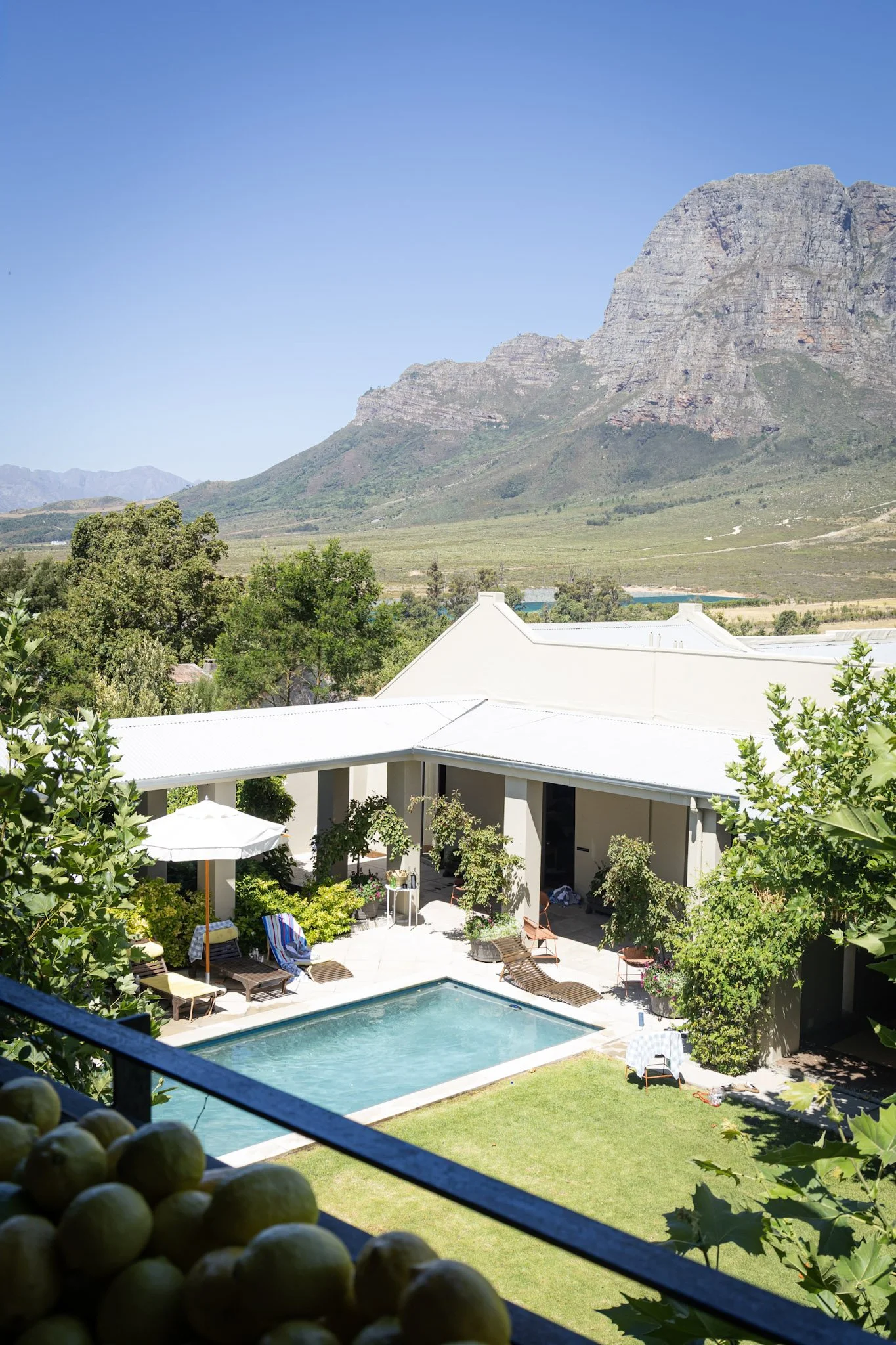 A backyard view with a swimming pool surrounded by lounge chairs, umbrellas, and green trees, with mountain ranges in the background and a clear blue sky above.
