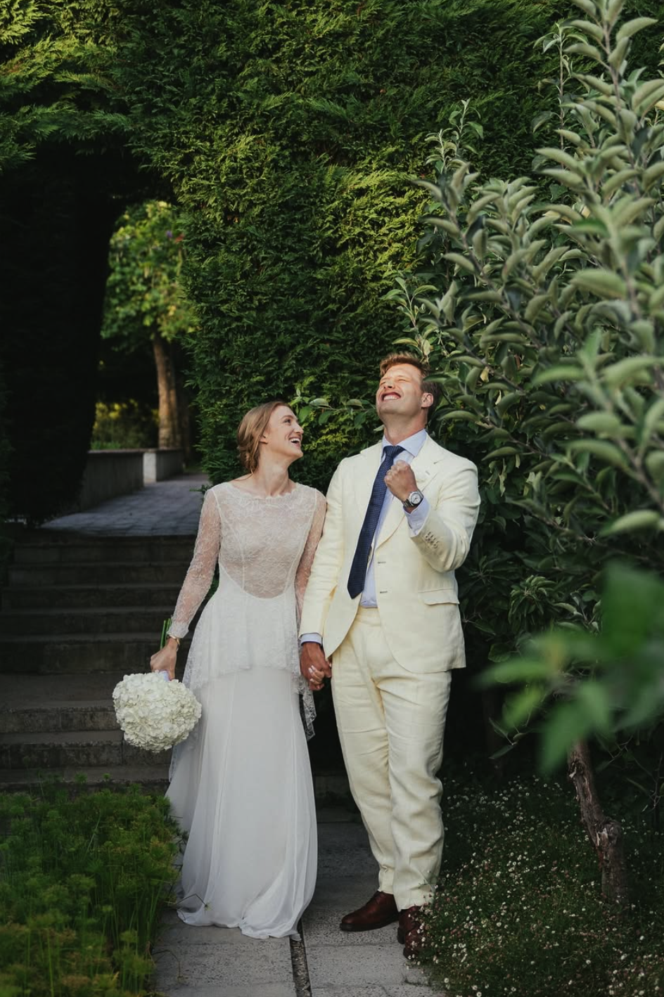 A wedding couple walking outdoors on a garden path, holding hands, surrounded by lush green trees and bushes, smiling joyfully.