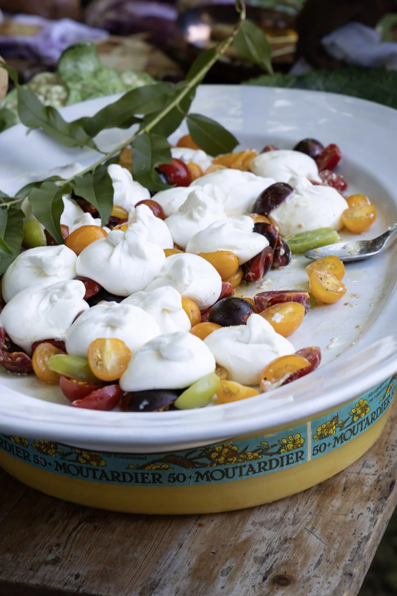 A round dish with a salad topped with dollops of mayonnaise or sour cream, cherry tomatoes, and green vegetables, with a green leafy branch laid across the dish. The dish is placed on a rustic wooden surface.