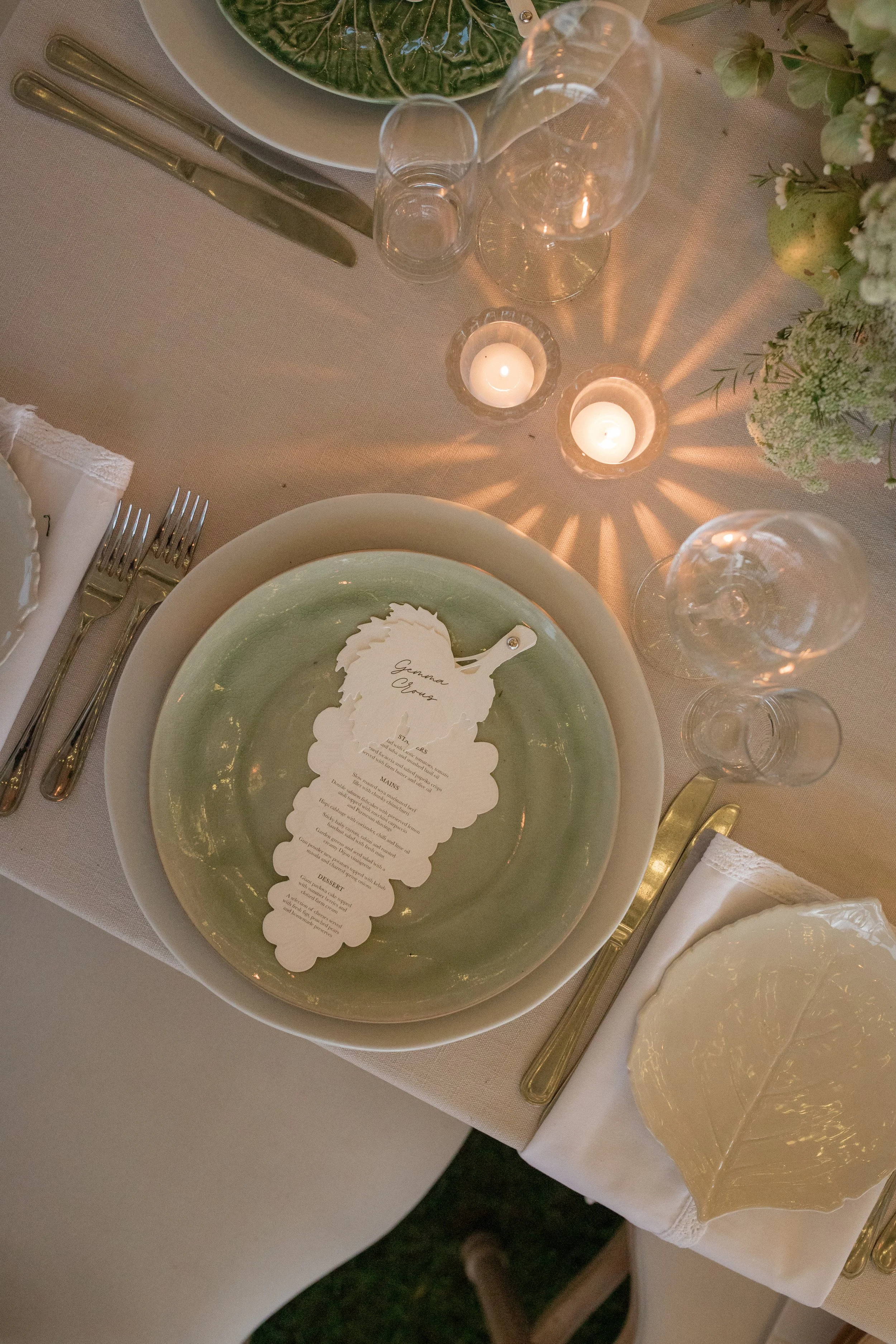 A table setting with a green and white plate, a menu, wine glasses, water glasses, candles, a napkin, and silverware, with floral decorations and soft lighting.