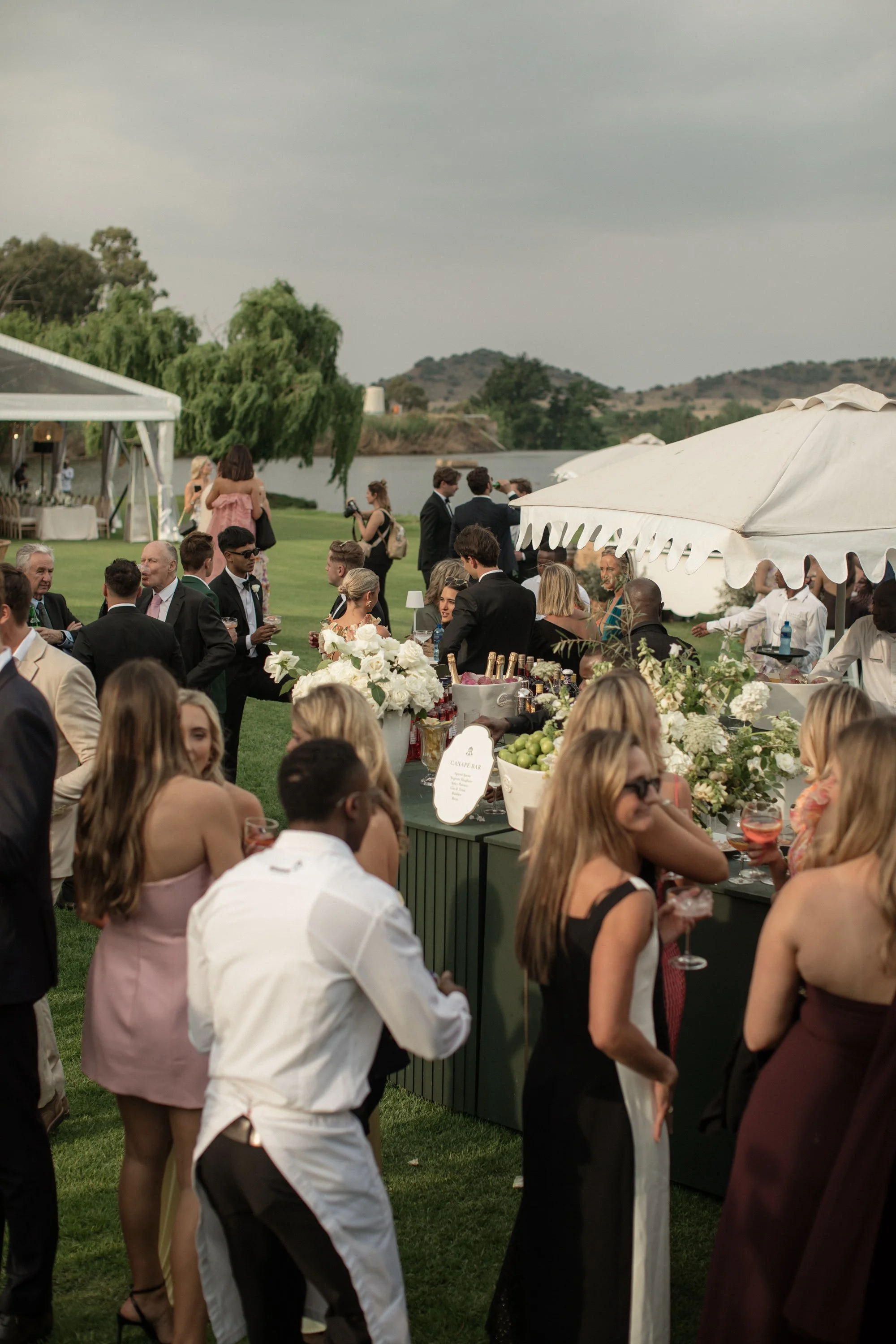 People gathered outdoors for a celebration, with some wearing formal attire, under a cloudy sky near water and hilly landscape, with a tent and decorative floral arrangements.