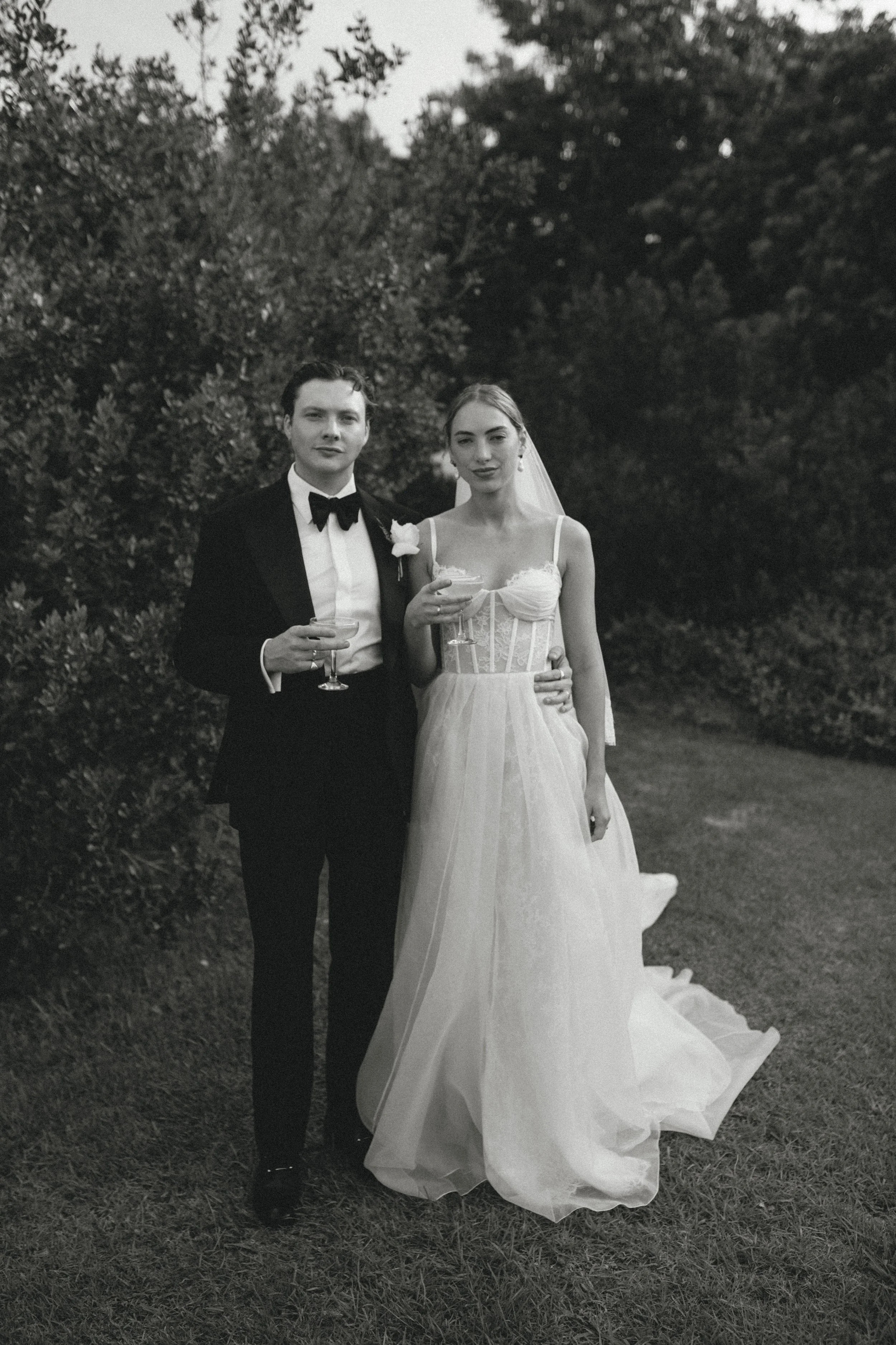 A black and white photograph of a newlywed couple standing outdoors in front of trees, dressed in wedding attire, holding drinks, with the bride in a white wedding gown and veil, and the groom in a tuxedo with a bow tie.