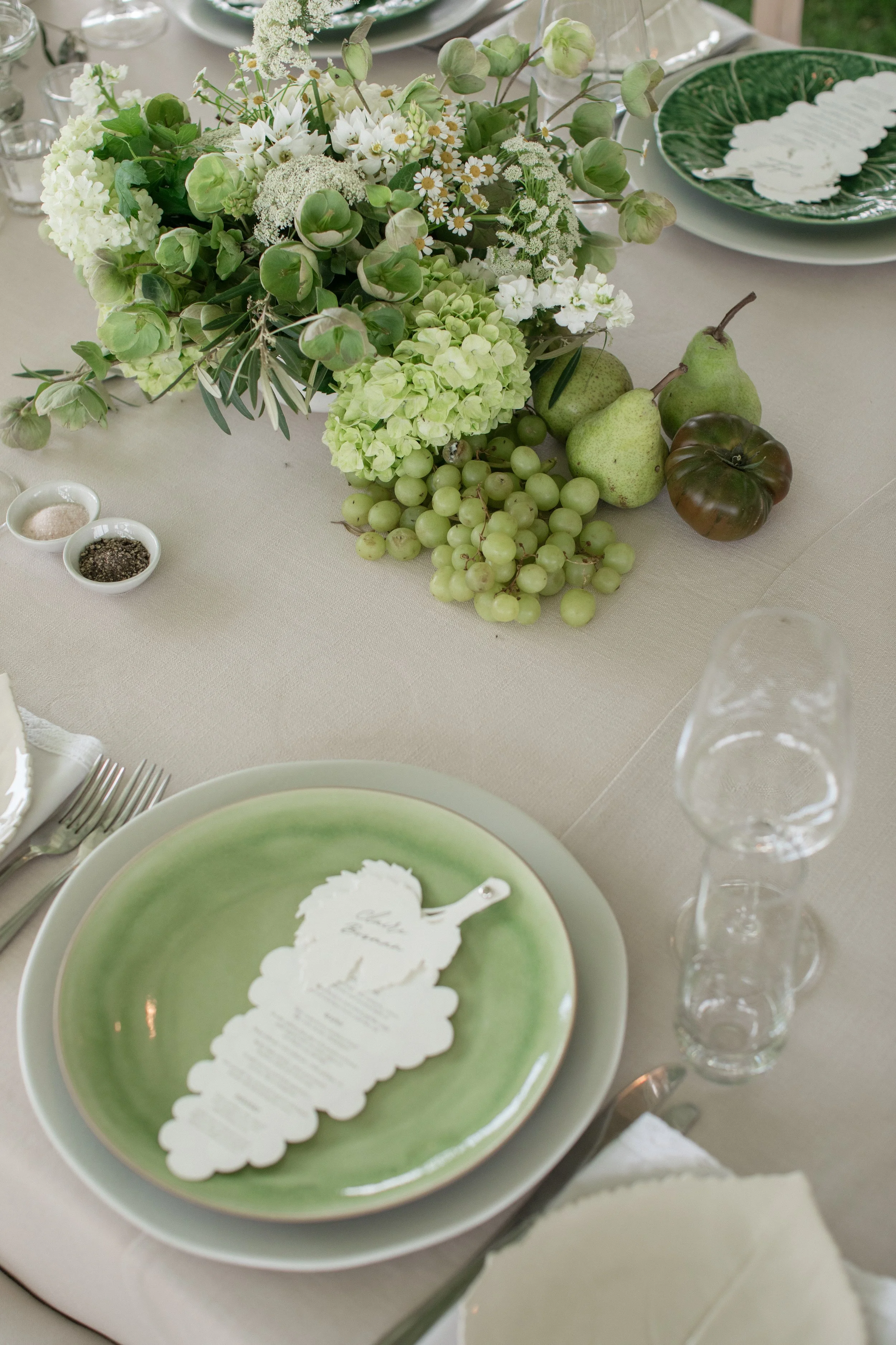 A table setting with a floral centerpiece, pears, grapes, a dark-colored tomato, a green and white plate with a paper cutout menu, and empty wine glasses.