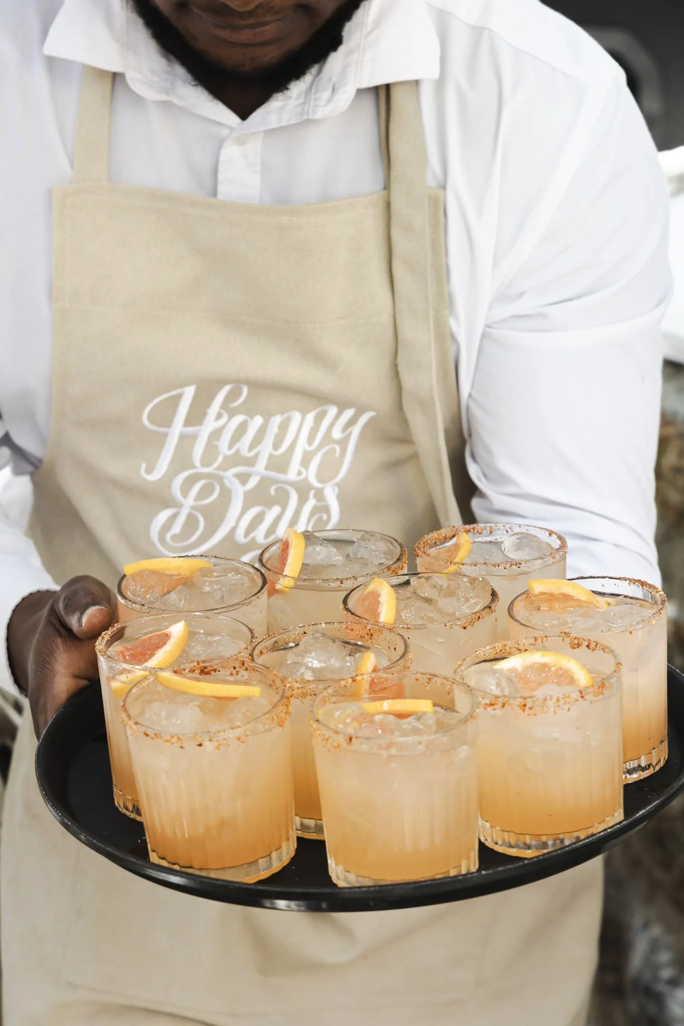 A person holding a tray of cocktails garnished with lemon slices and rimmed with chili powder, with a beige apron that says 'Happy Days' in white script.