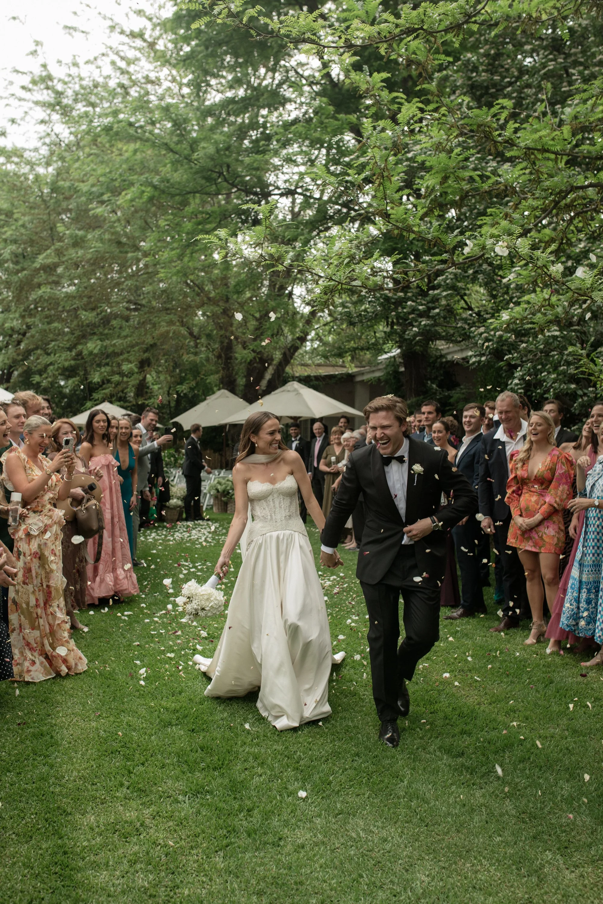 A bride and groom walking hand in hand under a green, tree-filled outdoor setting, surrounded by smiling wedding guests, some taking pictures, during a wedding celebration.