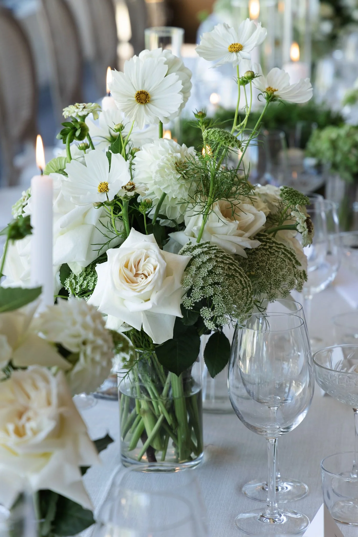 Close-up of a centerpiece with white flowers in a jar, surrounded by empty wine glasses on a table, with a blurred background of a decorated dining area.