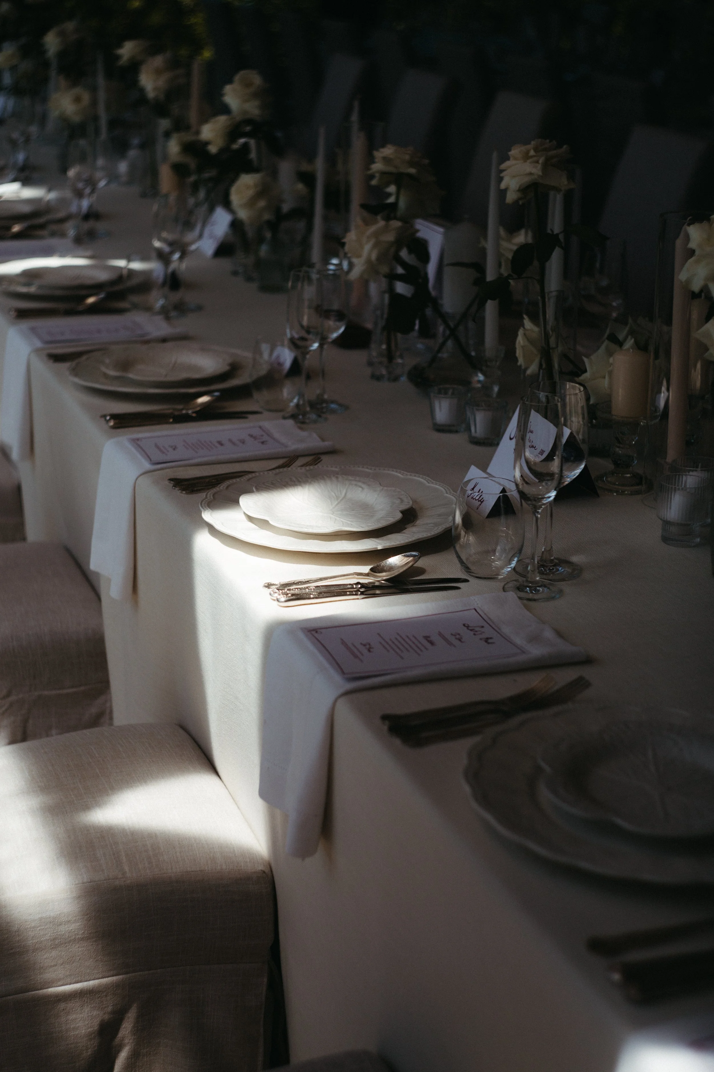 A long banquet table set for a formal event with white tablecloths, plates, utensils, wine glasses, and floral centerpieces with white flowers and candles.