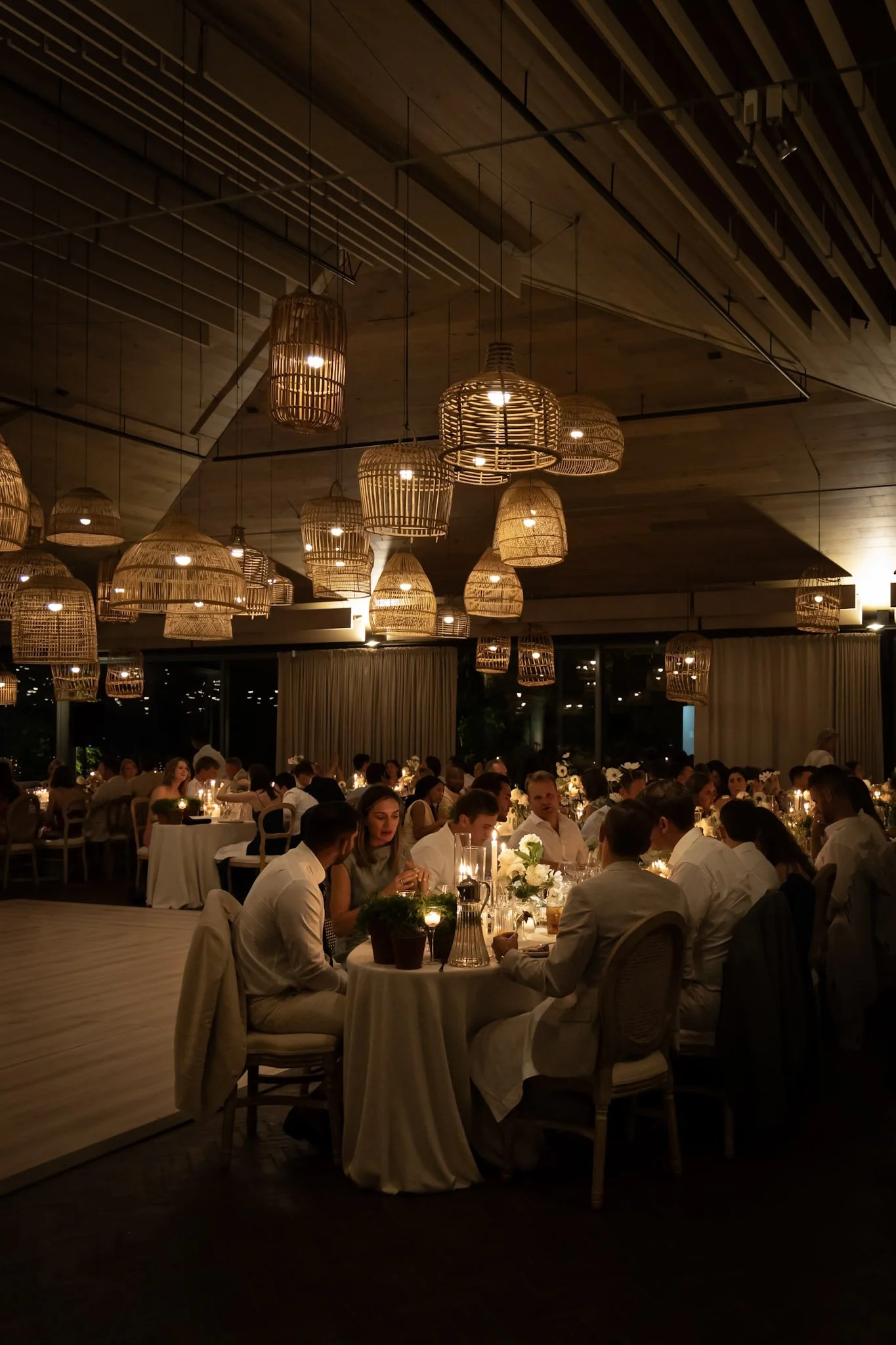 A large, elegant indoor dining area with warm lighting from woven pendant lamps hanging from a wooden ceiling. Guests in formal attire are seated at round tables with white tablecloths, floral centerpieces, and candlelit ambiance, enjoying a meal.