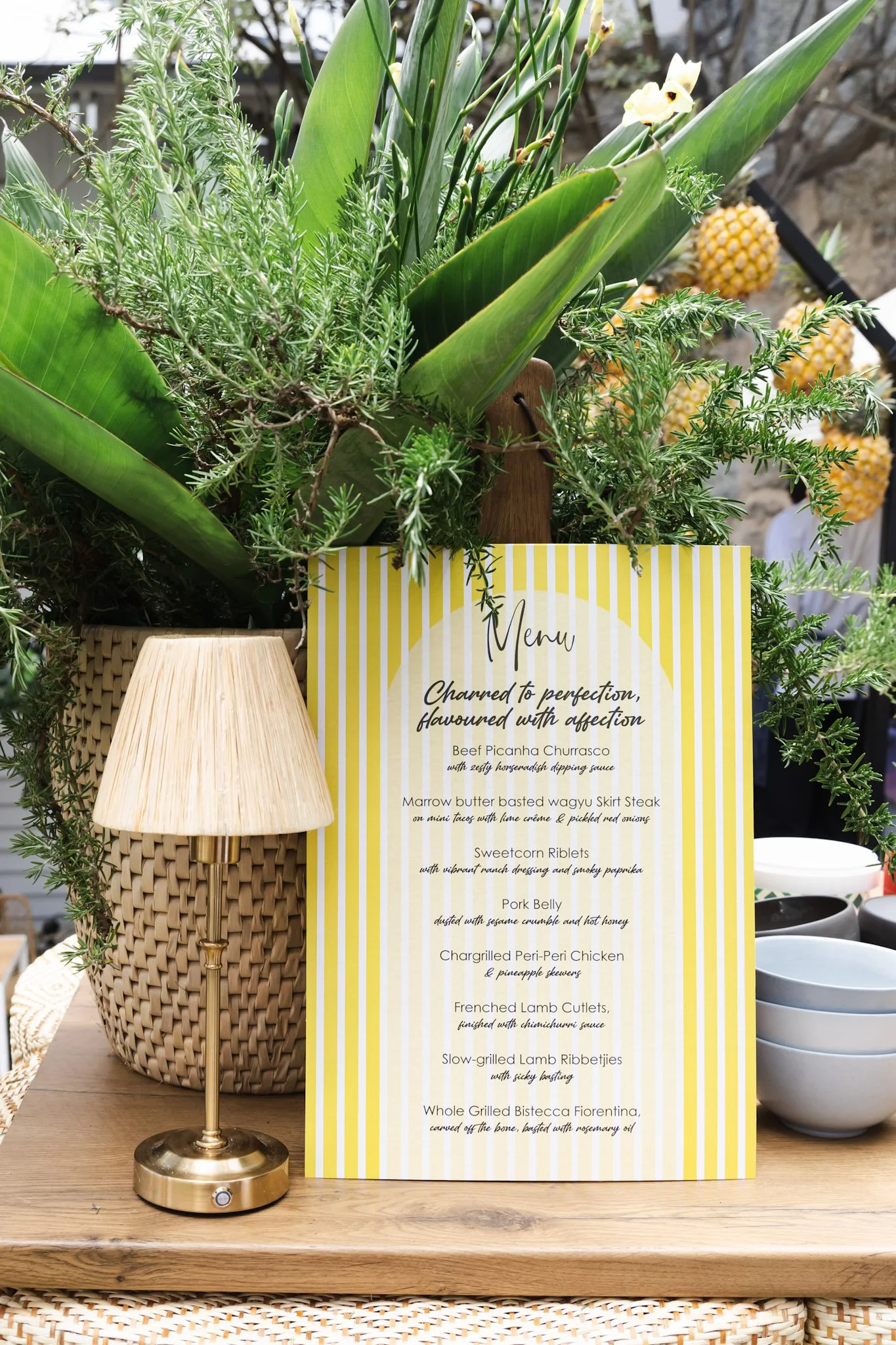 A table setting featuring a yellow striped menu with a list of dishes, a woven basket with greenery, a brass table lamp, and a background of pineapples and ceramic bowls.