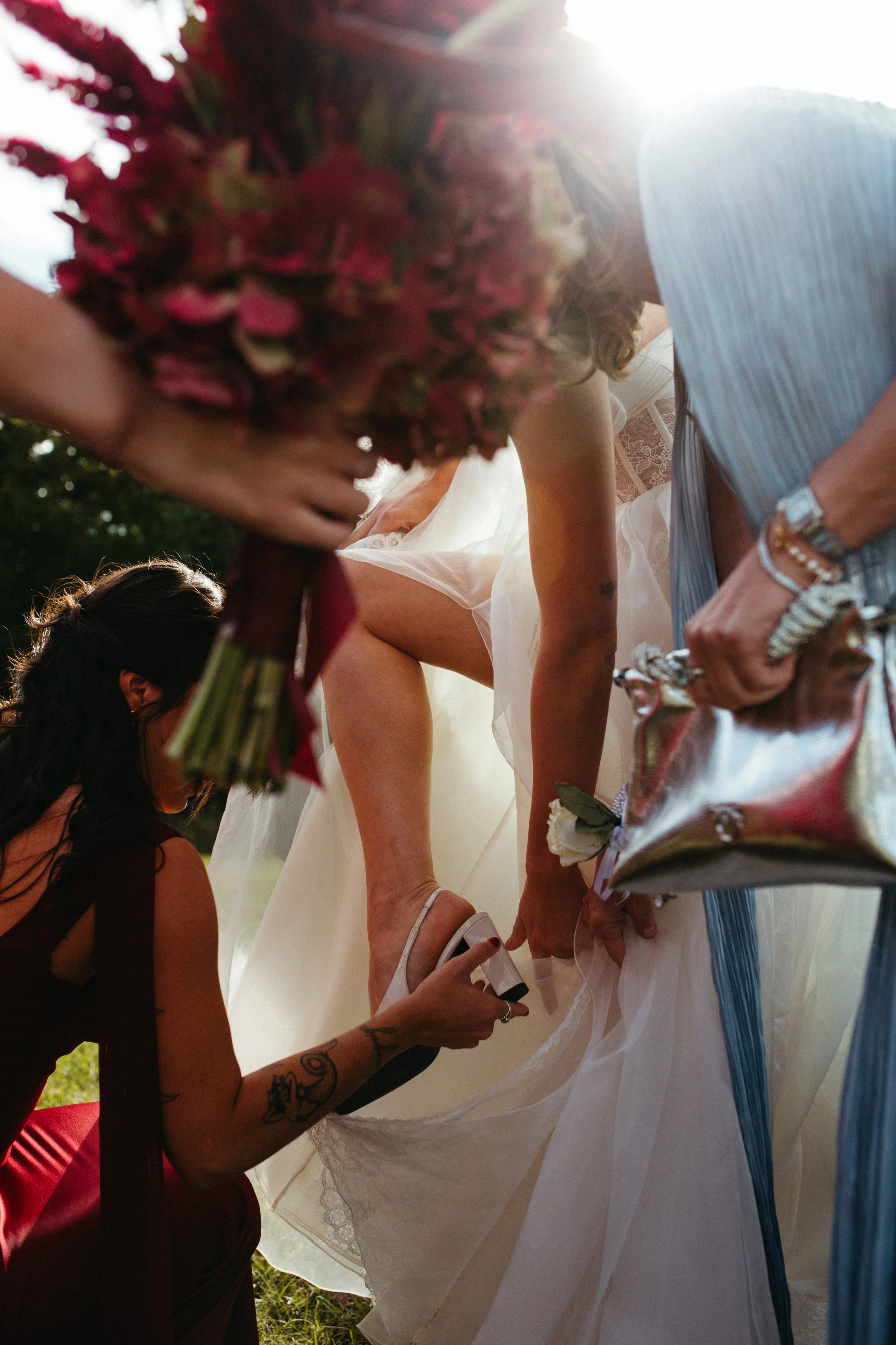 A woman getting her shoe fastened by another woman at a wedding or formal event, with floral bouquet held nearby, and sunlight in the background.
