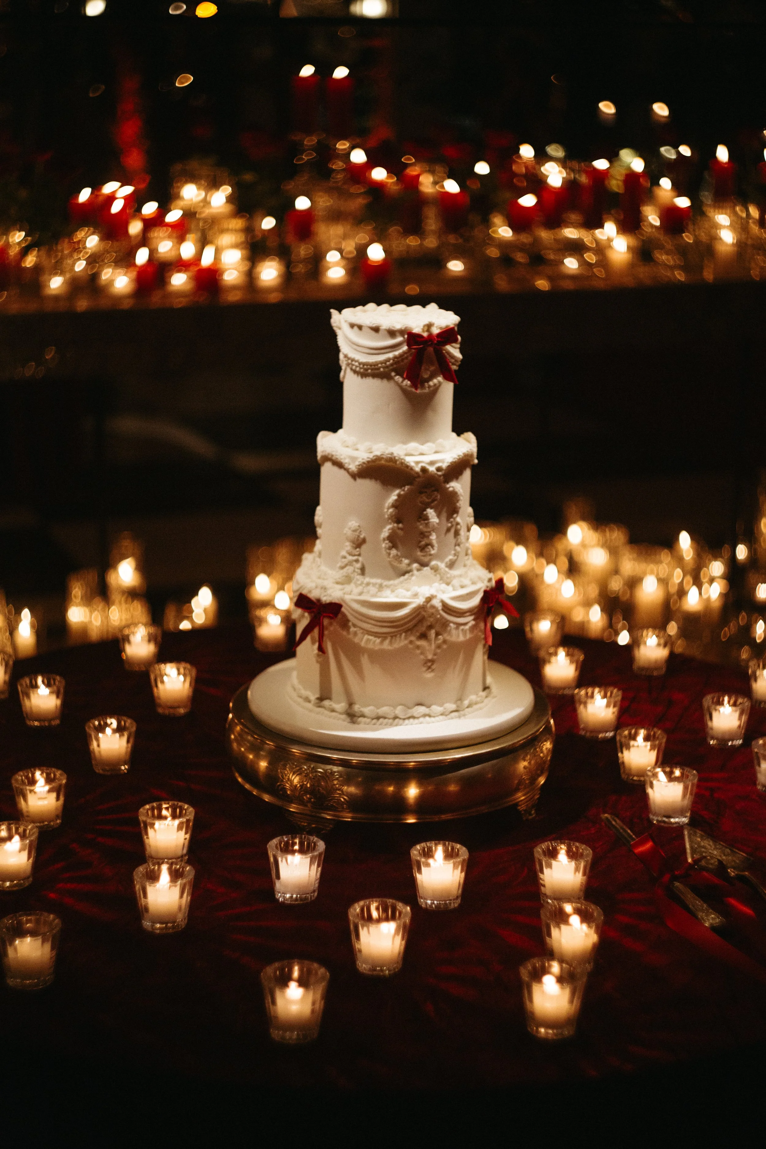A white wedding cake decorated with red bows and white icing, surrounded by numerous lit candles on a dark table.