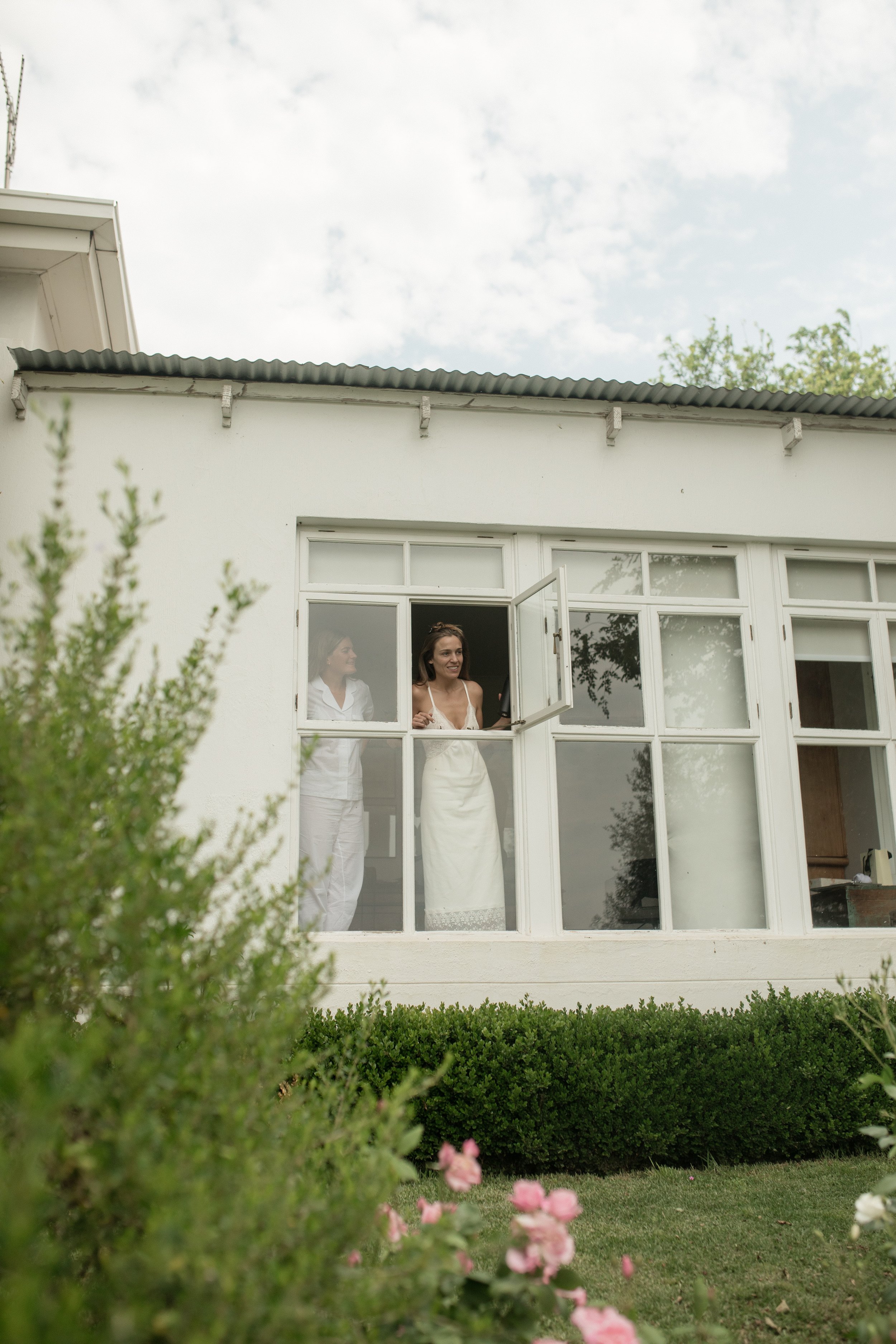 Two women in white dresses standing at the window of a white house, smiling and looking outside, surrounded by green bushes and plants.