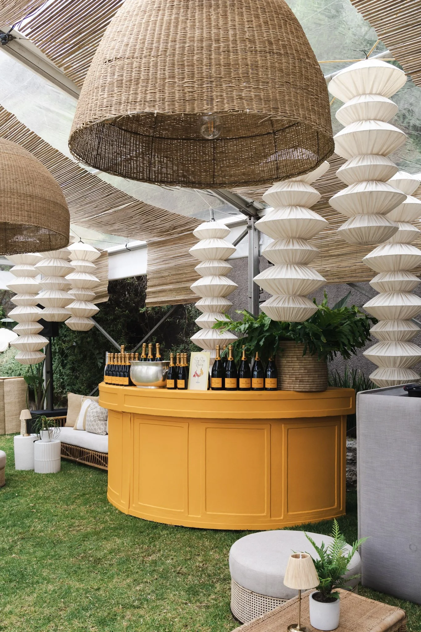 Decorative outdoor setup featuring a yellow bar on a grassy area, set with bottles and glasses, surrounded by lounge furniture, large woven light fixtures hanging from a ceiling, and lush greenery.