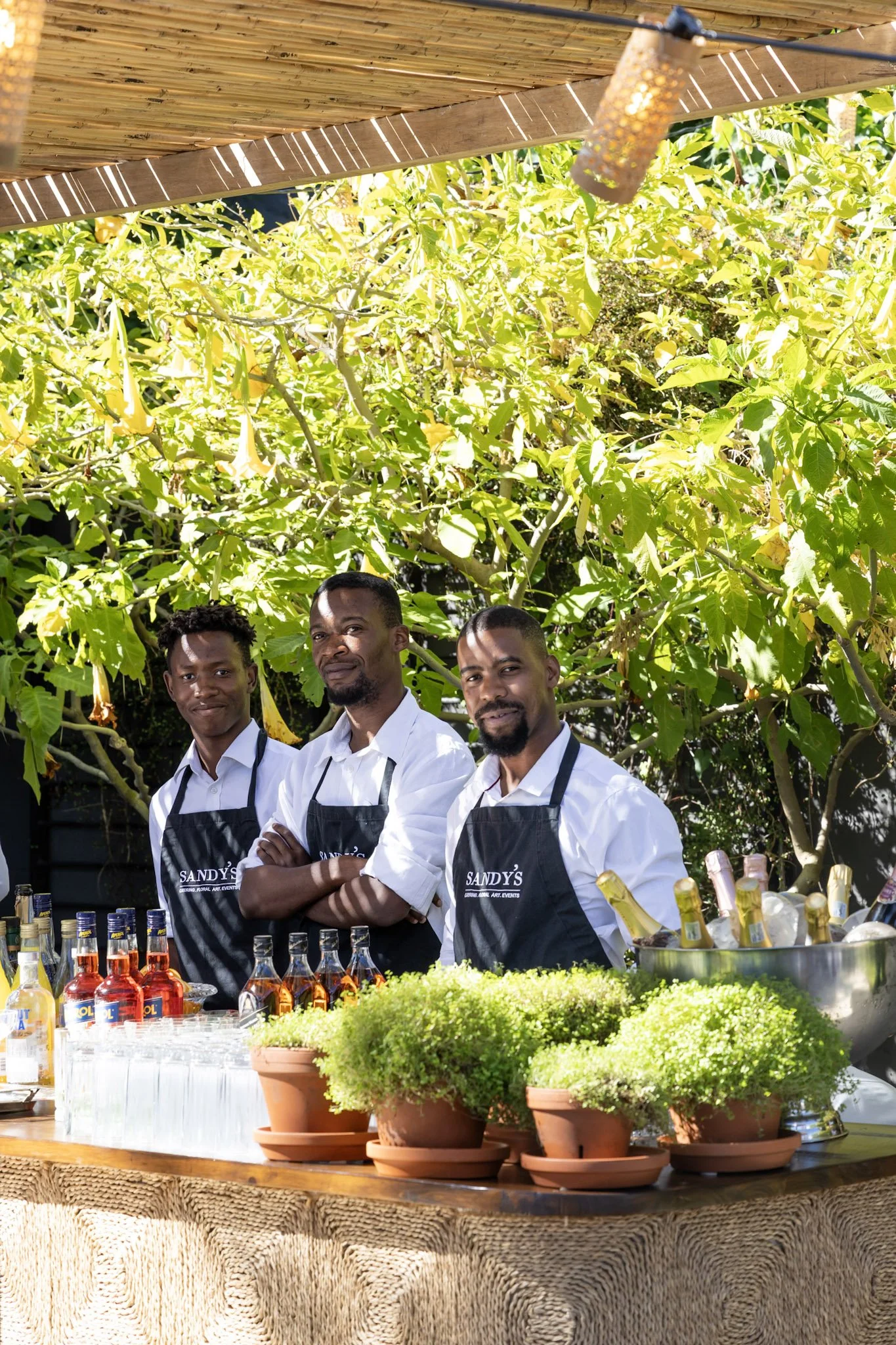 Three men wearing white shirts and black aprons with 'SANDY'S' logo standing behind a bar with potted plants and bottled drinks, outdoors under a bamboo canopy with leafy trees in the background.