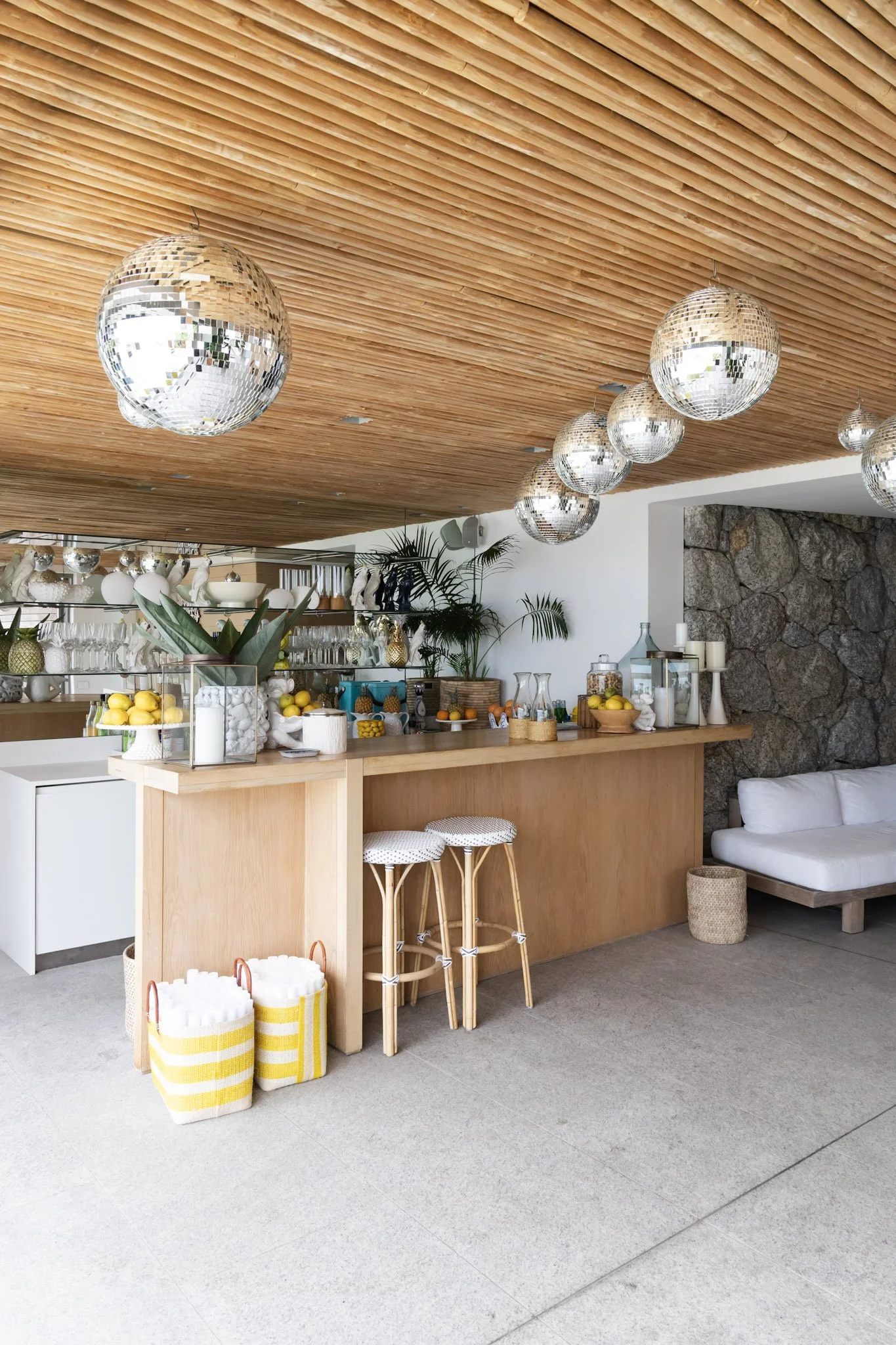 A modern indoor bar area with a wooden ceiling and disco ball lights, a stone wall, a white couch, and a wooden bar with various fruits, glasses, and decorative items.