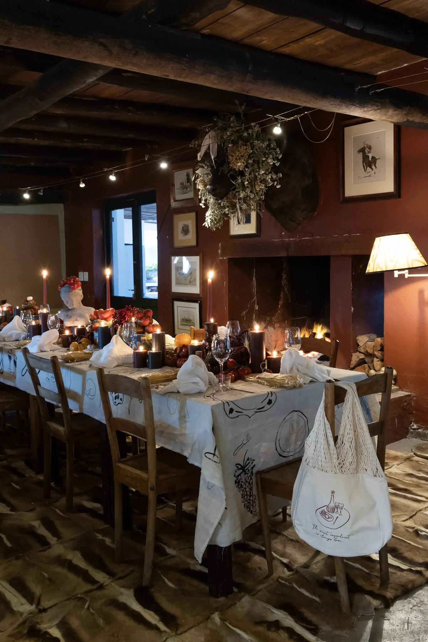 A cozy dining room with a long table set for a holiday meal, decorated with candles, fruits, and white napkins. A fireplace with fire is in the background, and there are framed pictures on the reddish-brown wall. The ceiling has exposed wooden beams,