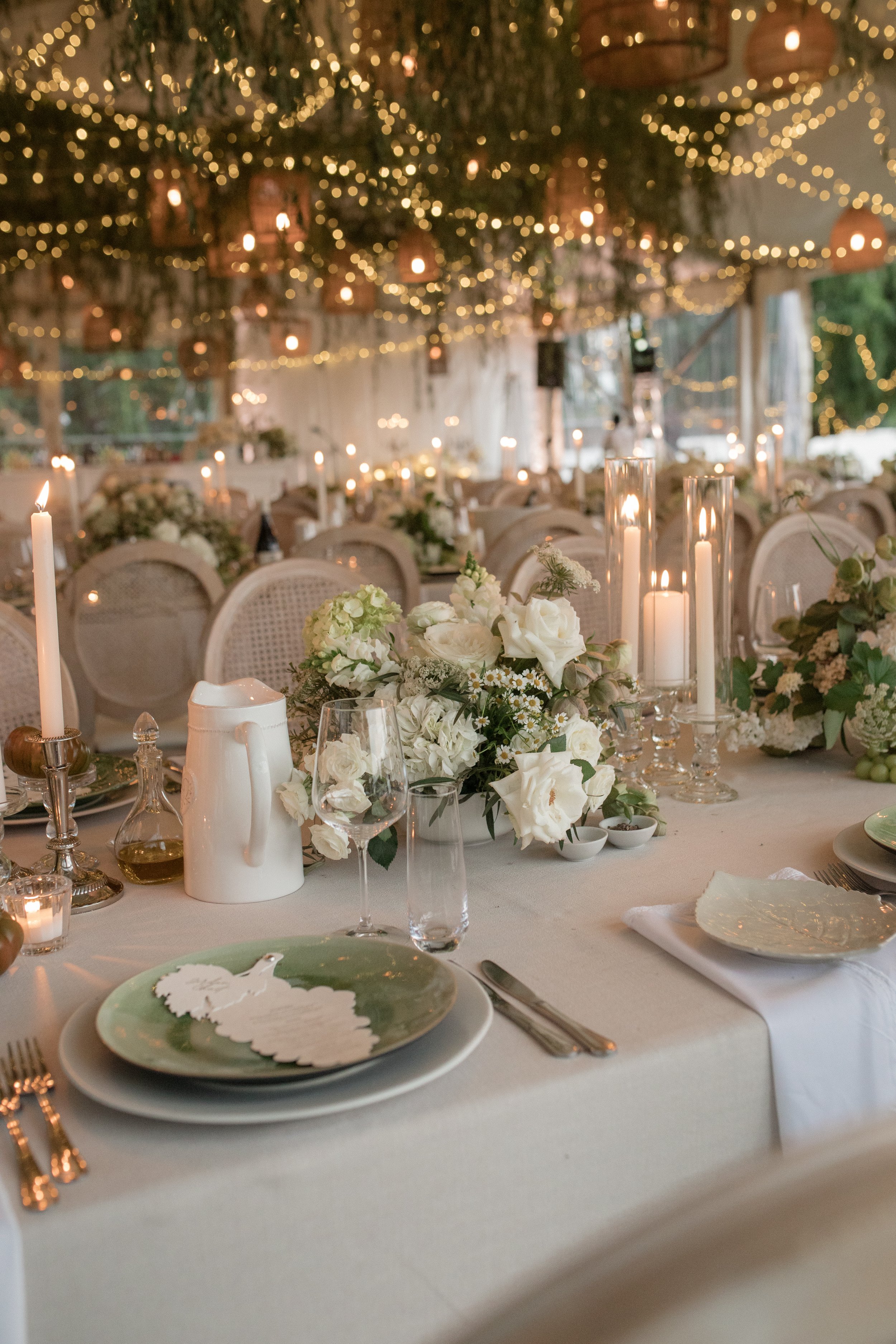 A decorated banquet table at an event with white floral centerpieces, tall candles, and wine glasses. In the background, there are string lights hanging from the ceiling, creating a warm ambiance.