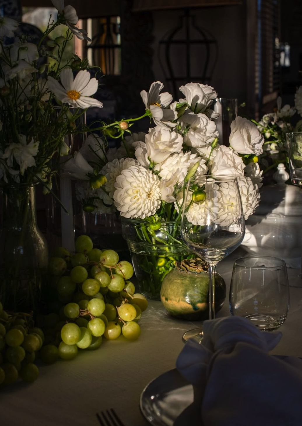 A table decorated with a variety of white flowers in vases, green grapes, and glassware, illuminated by natural light.