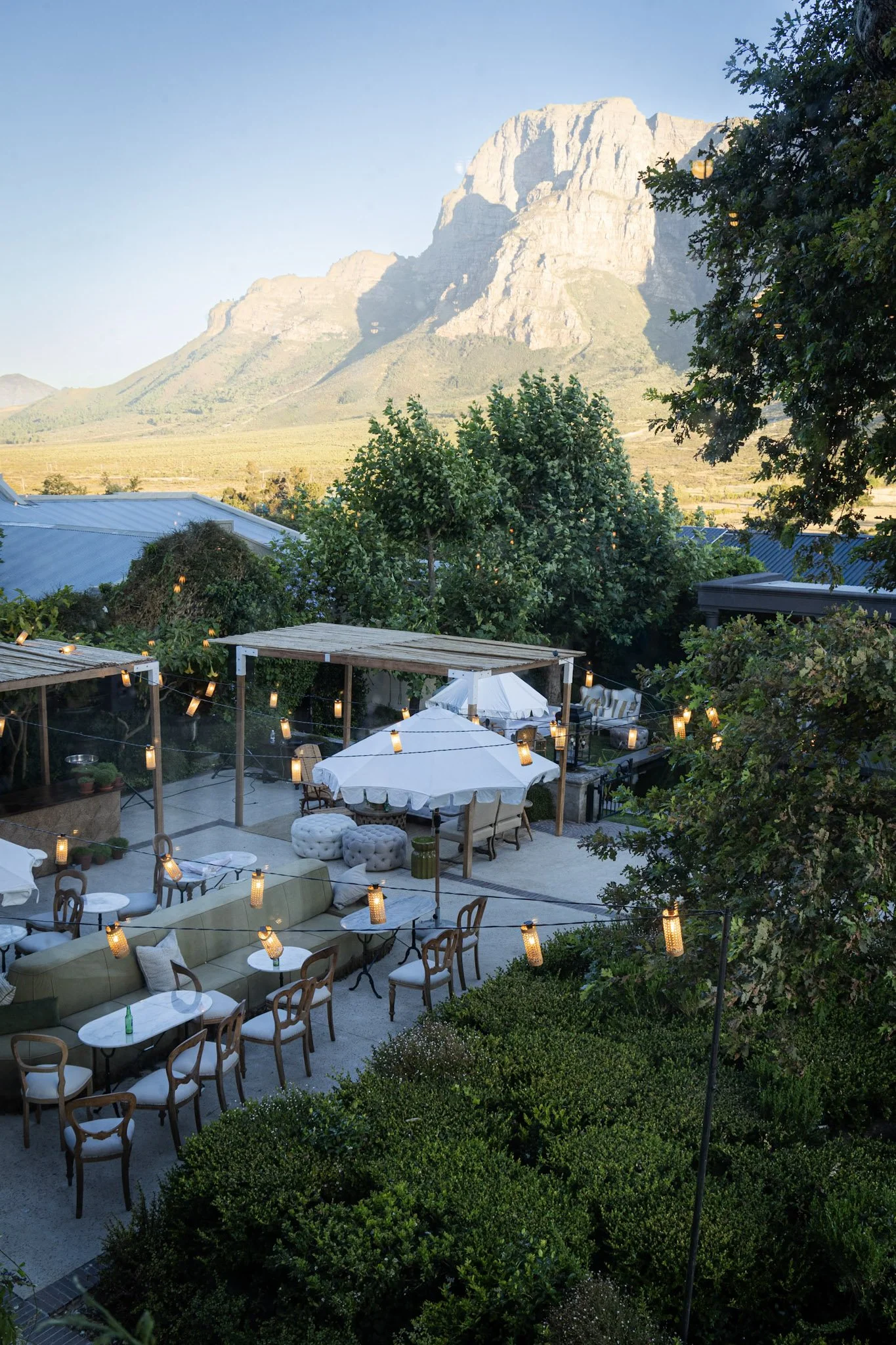 Outdoor restaurant or patio area with string lights, outdoor furniture, umbrellas, lush green trees, and a mountain in the background.