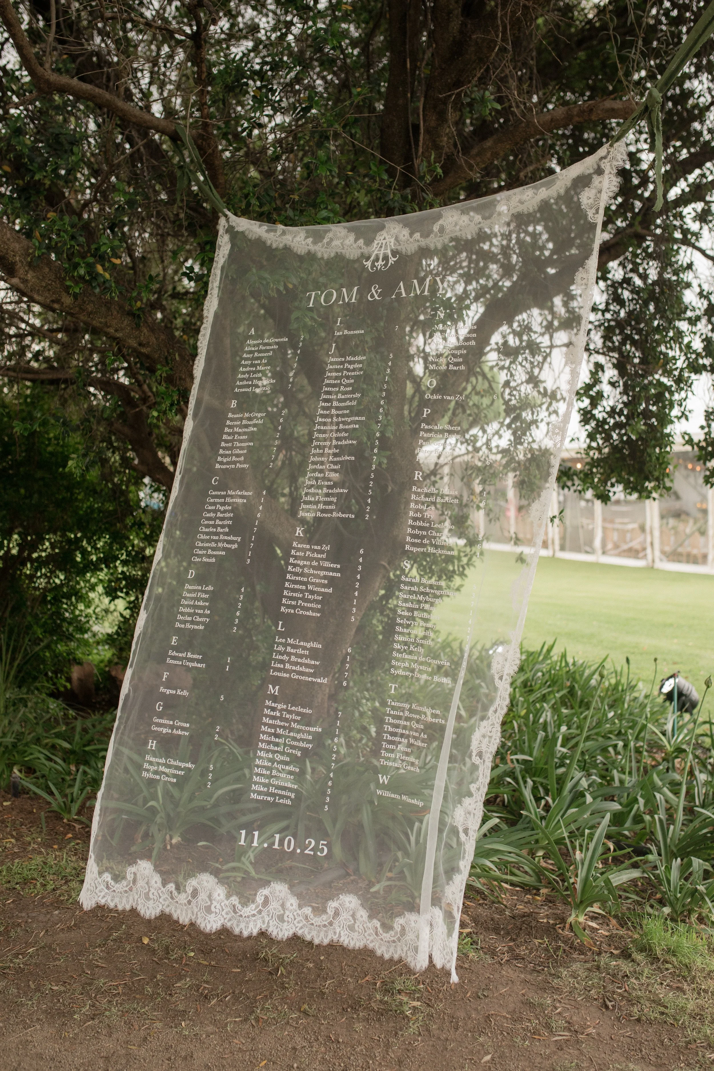 A transparent lace-edged fabric with embroidered text, hanging on a tree branch, displaying a list of names and numbers for a wedding seating chart, with trees and grass in the background.
